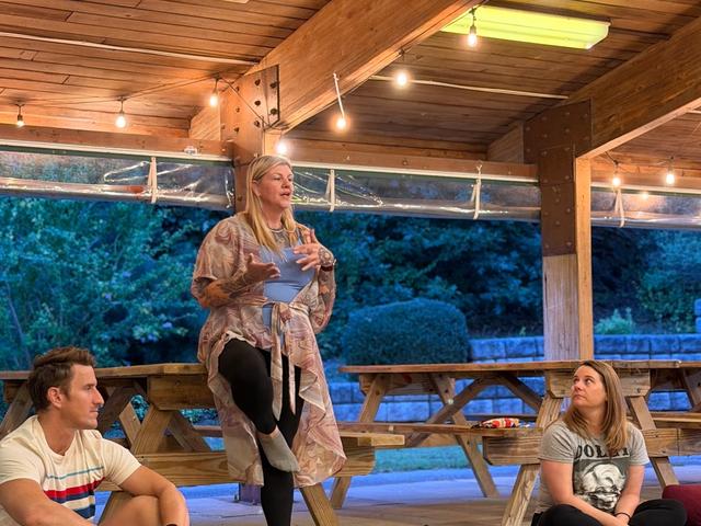 A woman standing and speaking to the group, who are seated on mats around her.