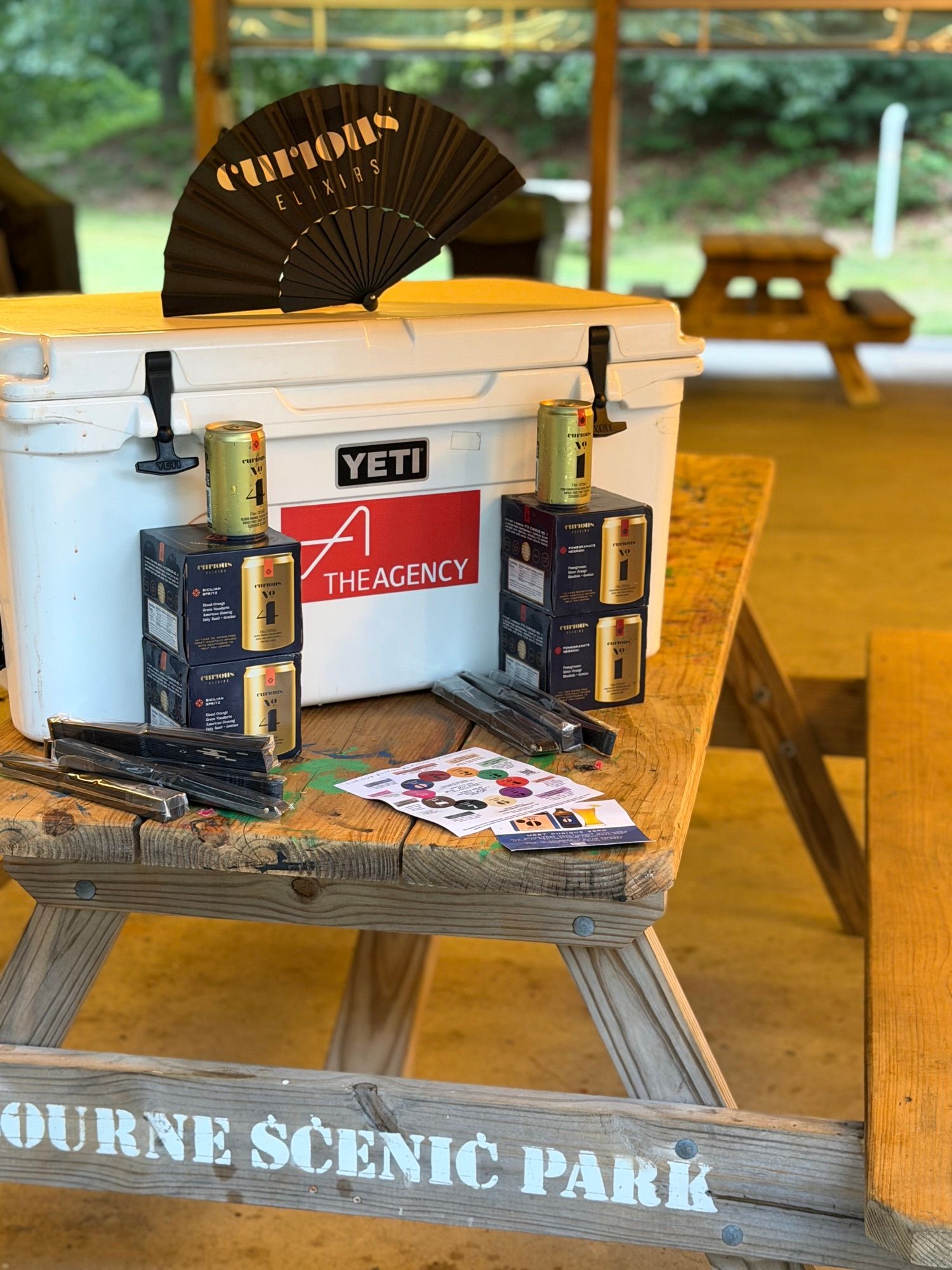 A cooler and boxes of Curious Elixirs set out on a picnic table stencilled “Bourne Scenic Park”.