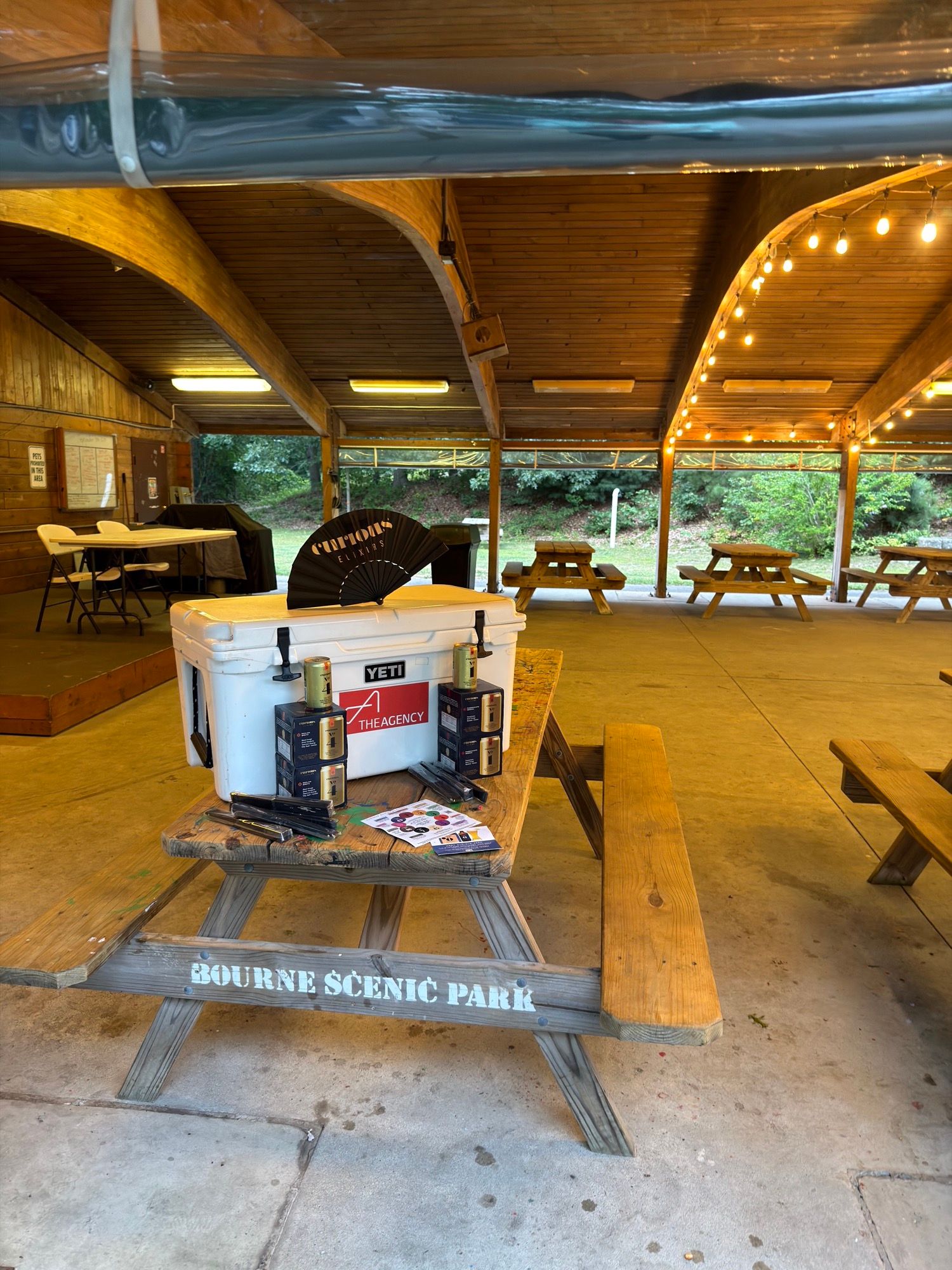 The open-sided wooden pavilion at dusk, string lights along the rafters and picnic tables around the edges.