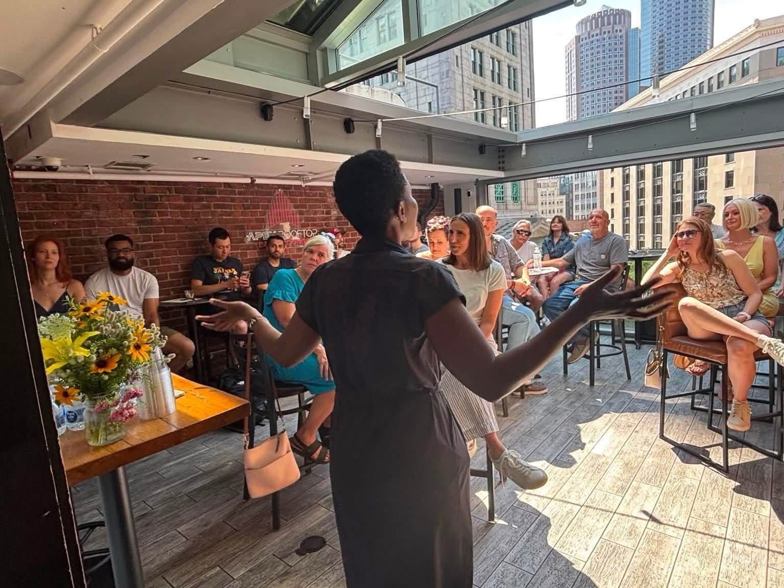 Woman with her back to the camera speaking to a seated crowd with the Boston skyline in the background.