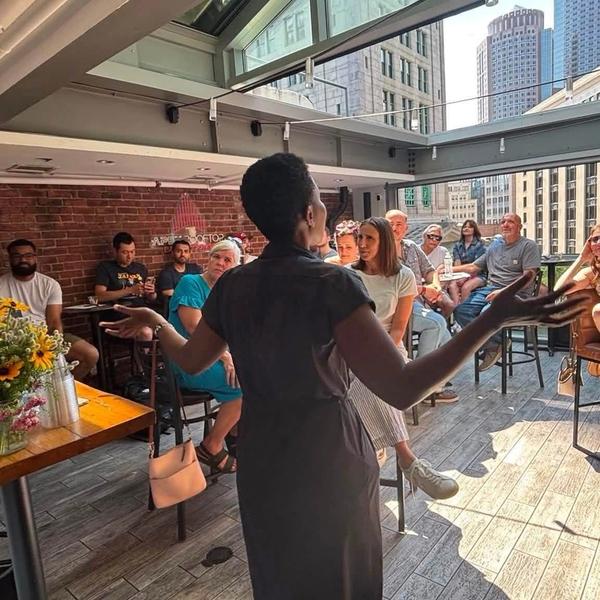 Woman with her back to the camera speaking to a seated crowd with the Boston skyline in the background.