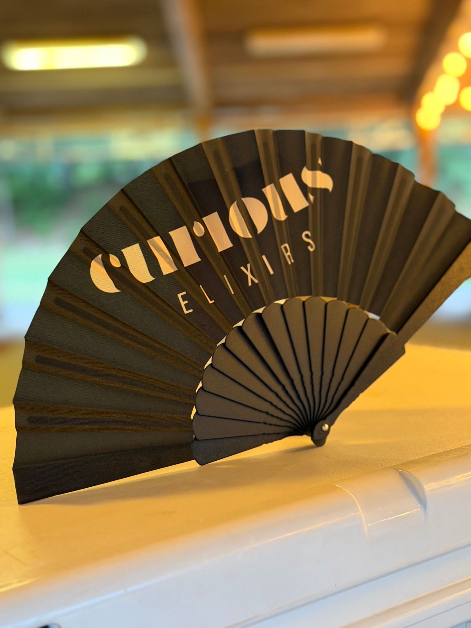 A black folding fan printed with the Curious Elixirs logo, propped on a cooler under warm string lights.