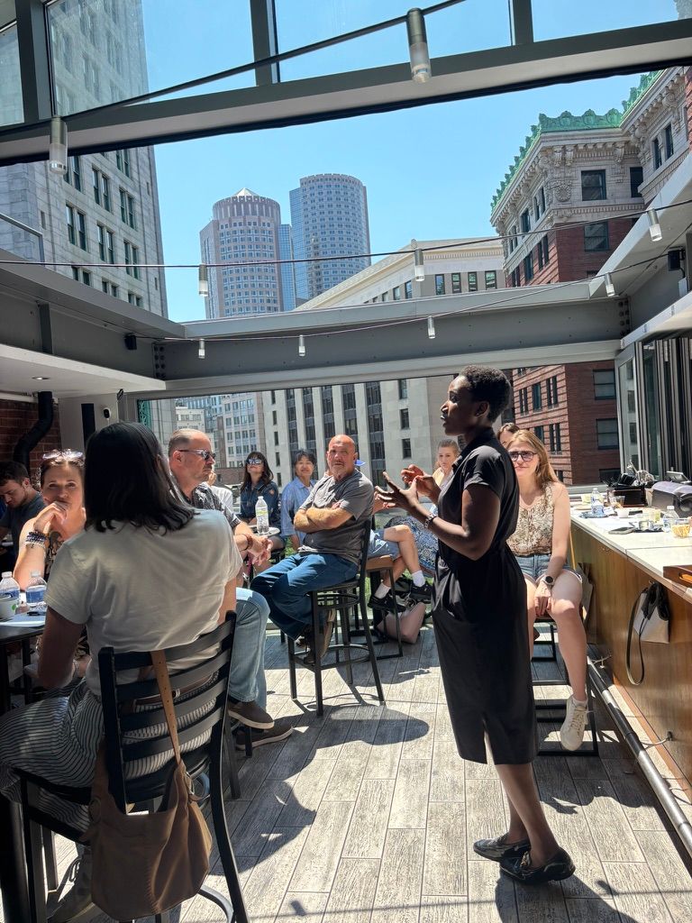 A host speaking to a seated group on a rooftop with the Boston skyline behind her