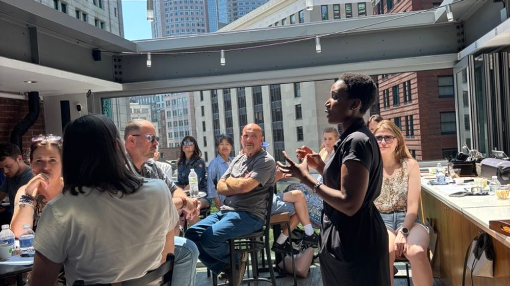 A host speaking to a seated group on a rooftop with the Boston skyline behind her