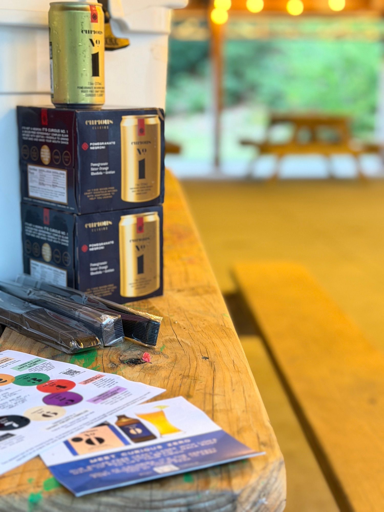 Boxes and a bottle of Curious Elixirs stacked on a picnic table beside a colourful information card.