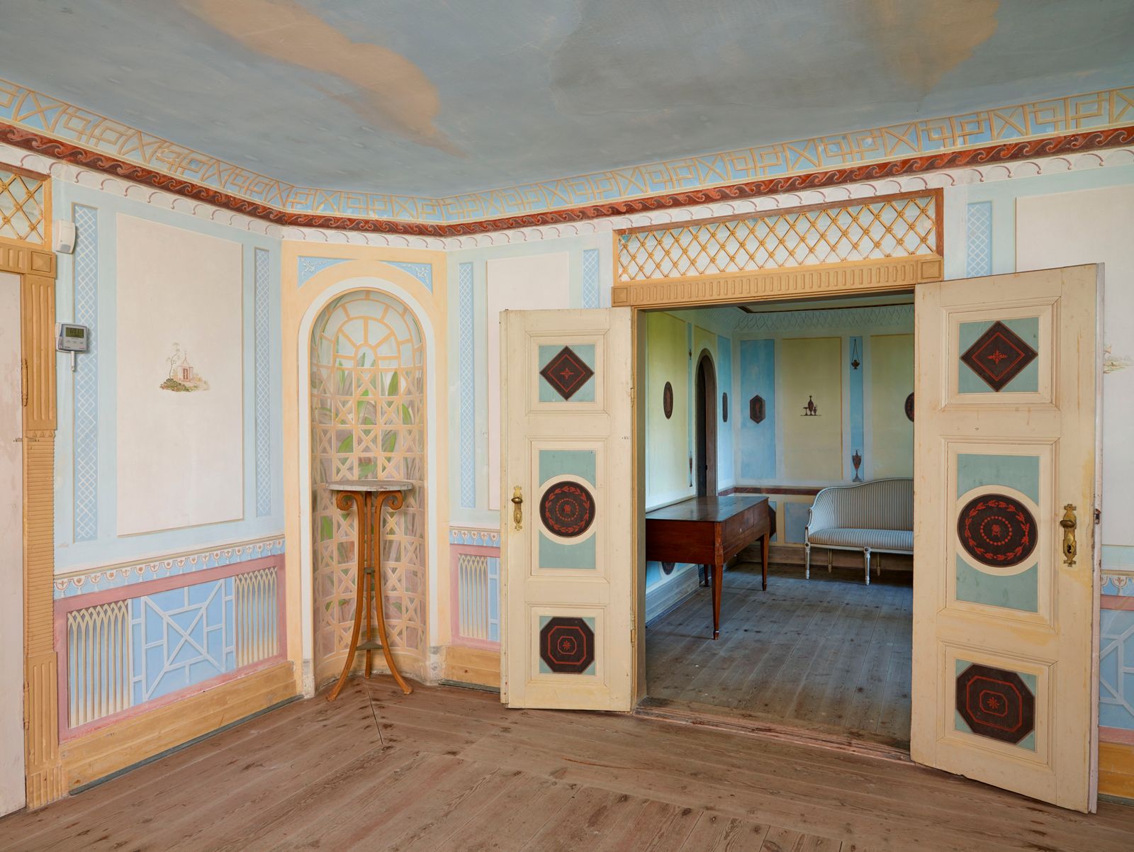 Den kinesiske stue i Det Norske Hus ved Liselund med farverigt dekorerede døre, der åbner til en tilstødende stue med bord og sofa.