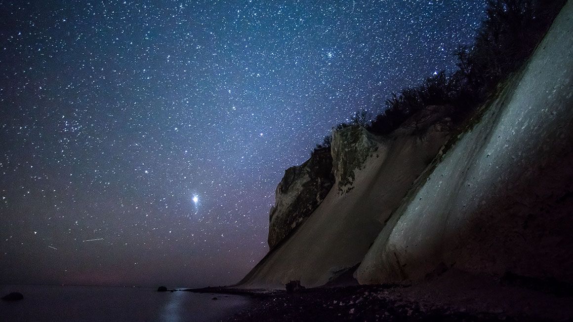 Møns Klint badet i stjernehimmel om natten, med havet og de hvide kridtklinter svagt oplyst af måneskin.