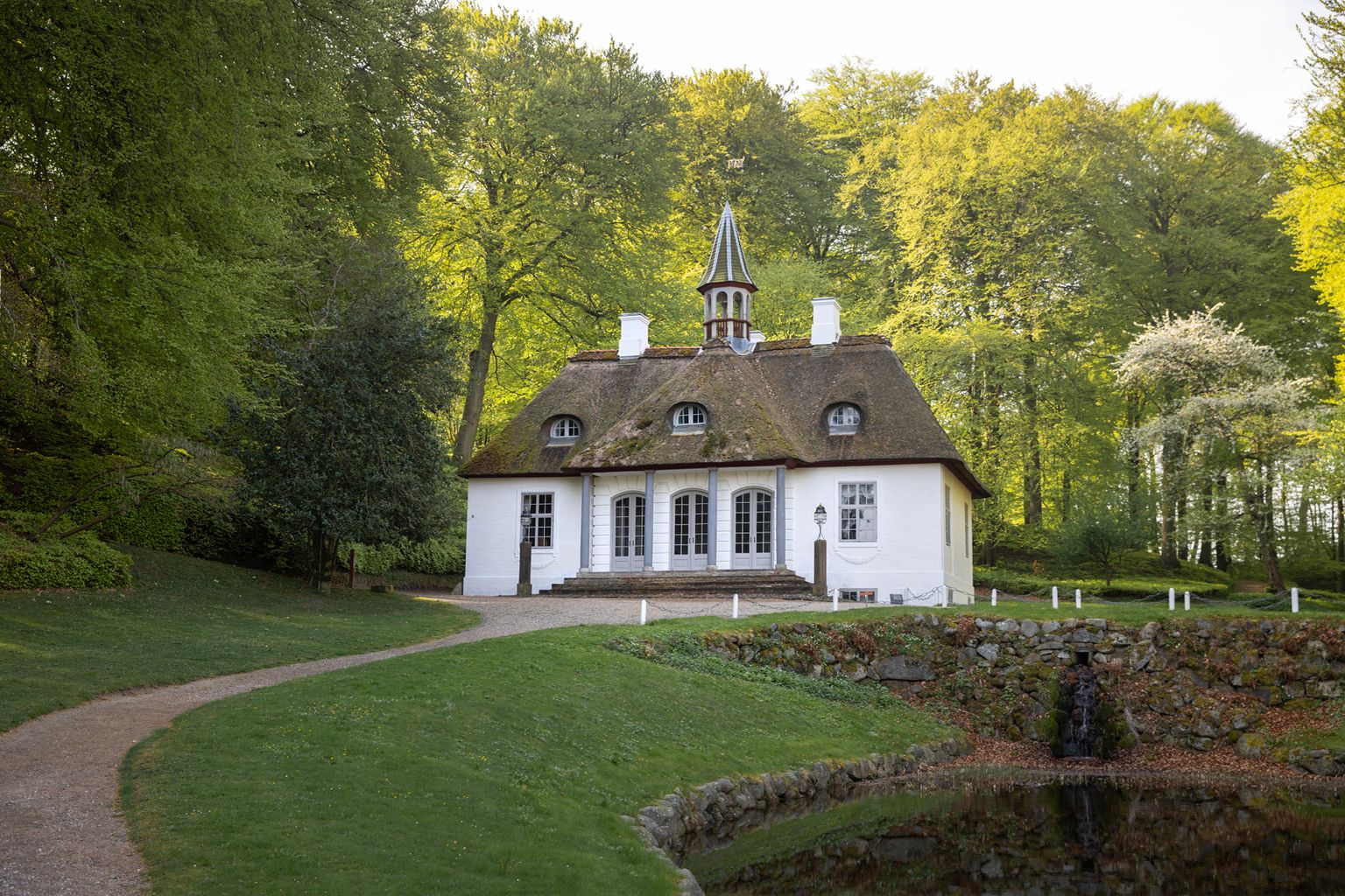 Forsiden af Liselund Lystslot med stråtag, hvide søjler og dørpartier, omgivet af nyudsprunget park om foråret.