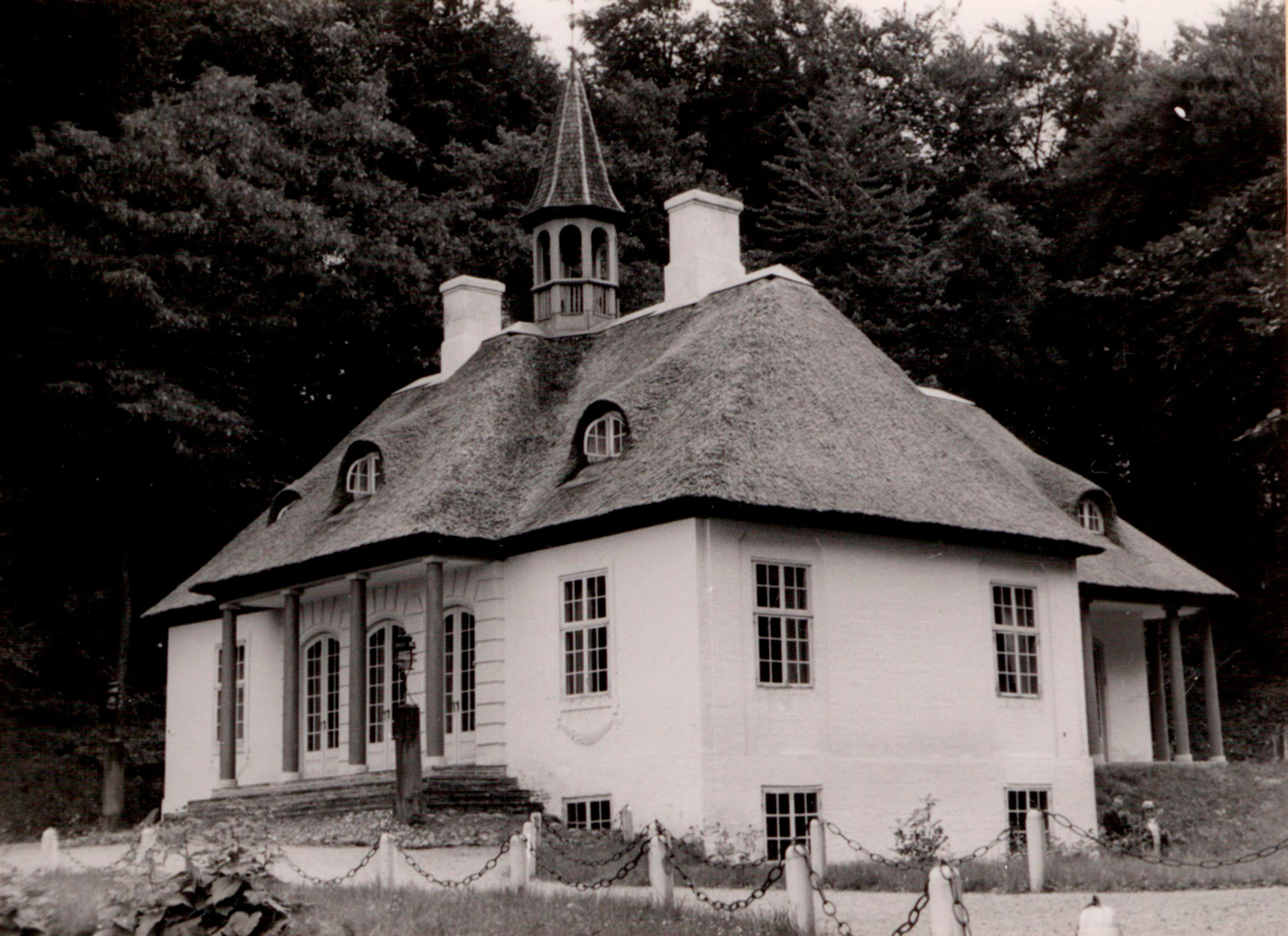 Historisk sort-hvidt fotografi af Liselund Lystslot med stråtækt tag, hvide facader og søjlegang, omgivet af høje træer.