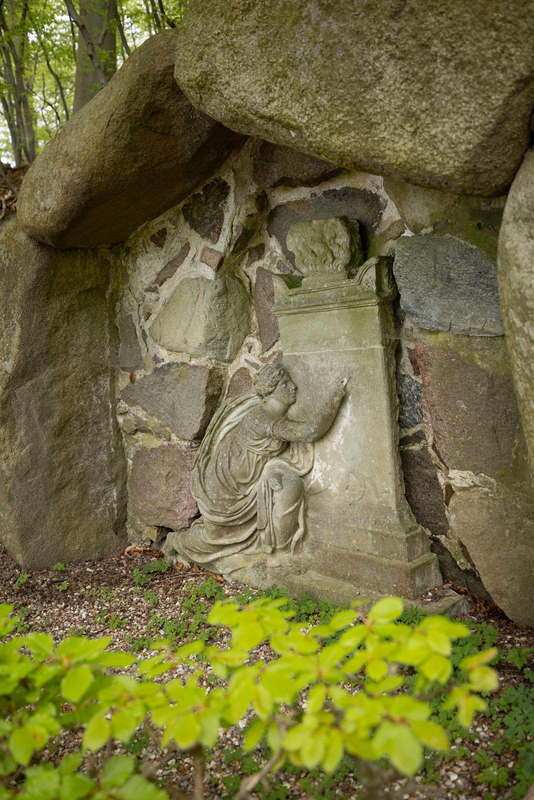 Stenrelief i en grotte i Liselund Park, der viser en knælende skikkelse foran et postament med en buste.