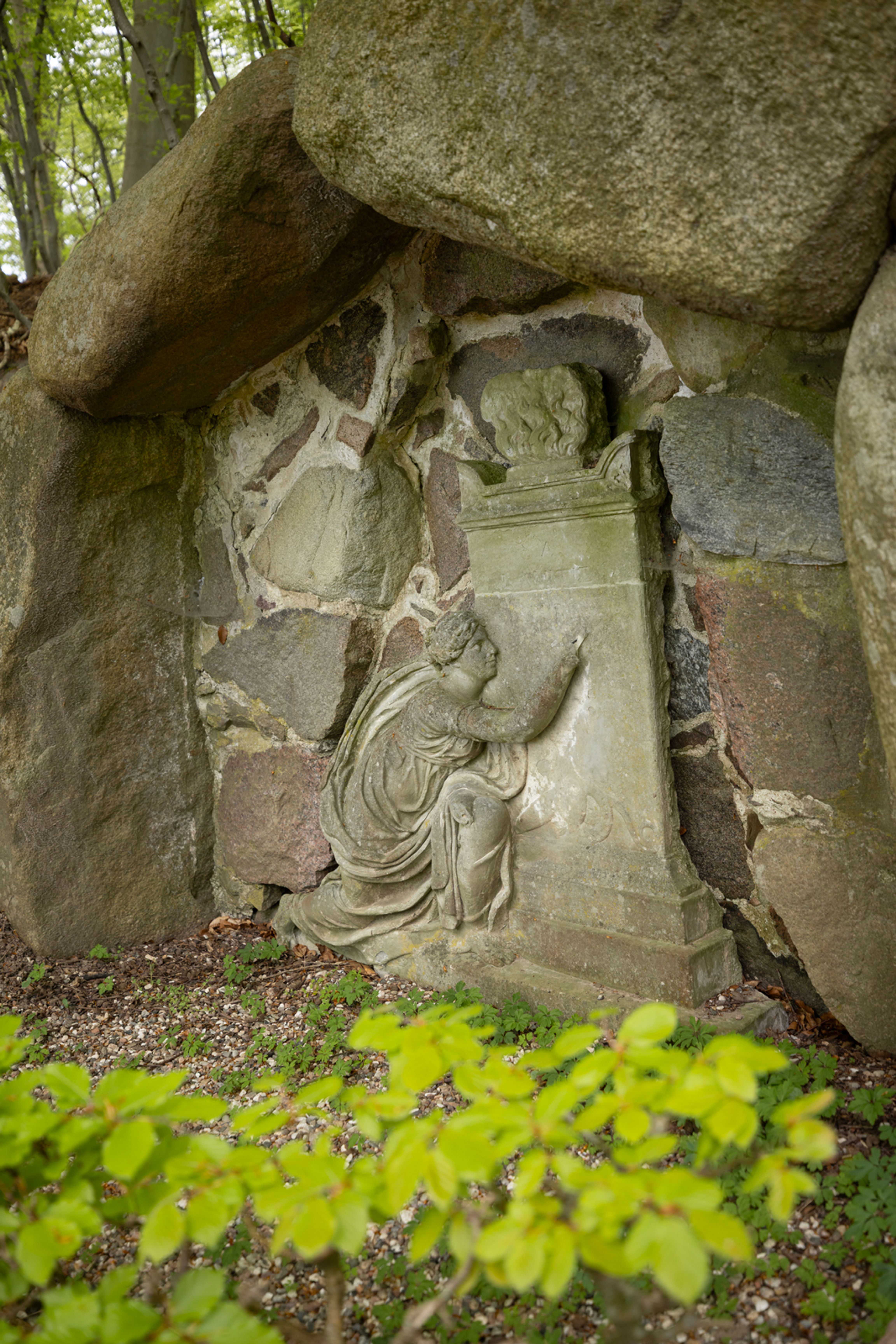 Stenrelief i en grotte i Liselund Park, der viser en knælende skikkelse foran et postament med en buste.