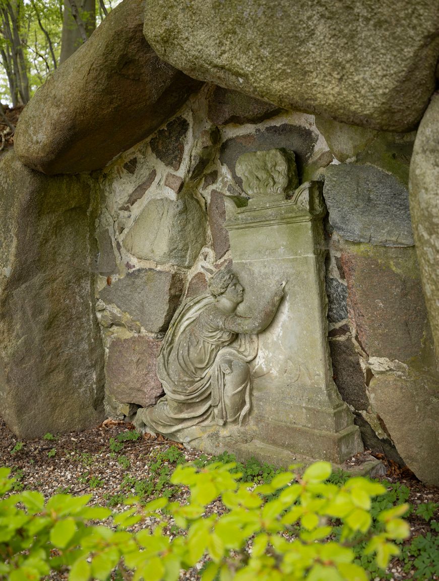 Stenrelief i en grotte i Liselund Park, der viser en knælende skikkelse foran et postament med en buste.