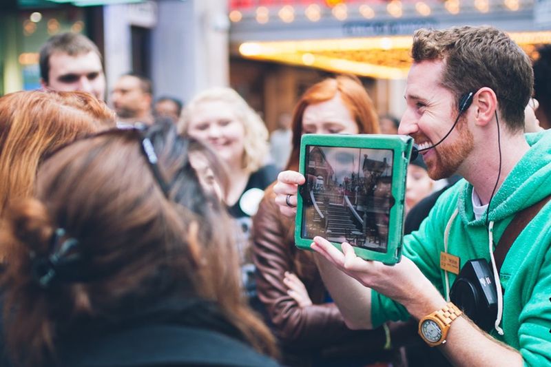 A Broadway Up Close tour guide showing a historical photo on a tablet to a small group in Times Square