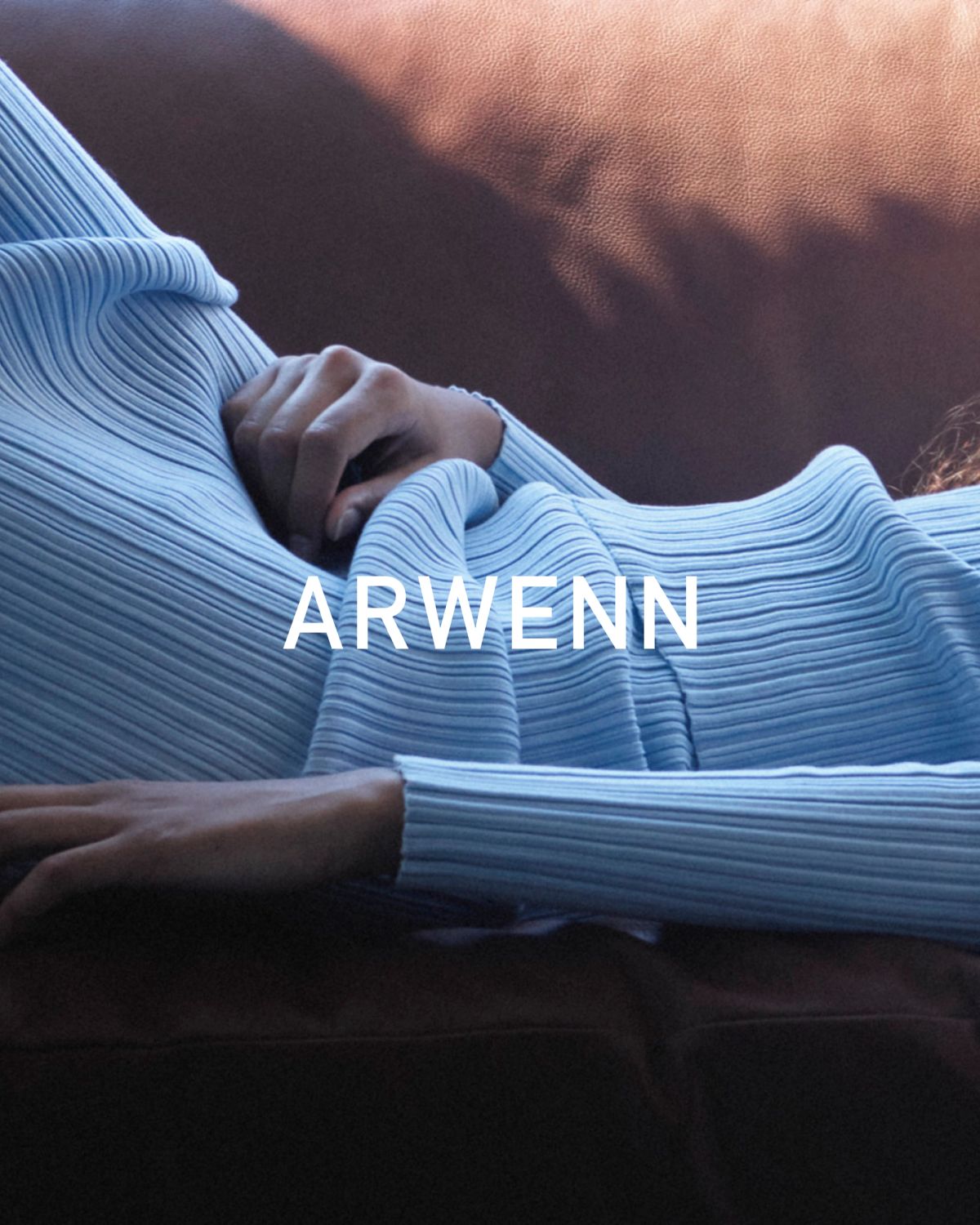 Close-up of a person in a light blue ribbed knit top lying on a brown leather sofa, one hand resting on their chest, soft light falling across the leather.