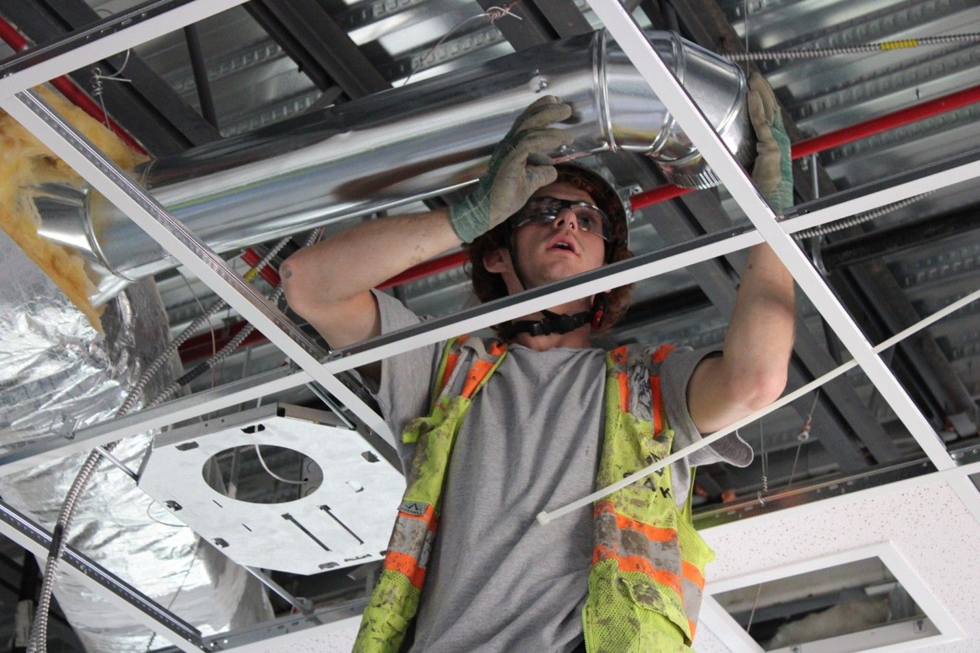 Technician installing commercial HVAC ductwork above a suspended ceiling while wearing a hard hat, safety glasses, gloves, and a high-visibility vest during a commercial construction project.