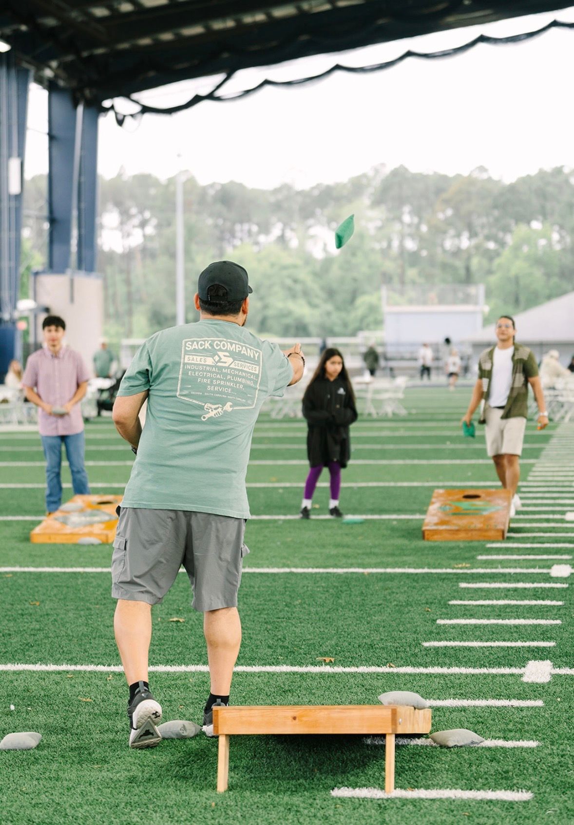 Employee playing cornhole at Sack Day on a turf field while coworkers watch in the background.