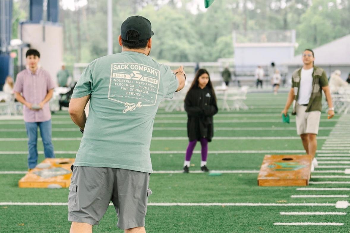 Employee playing cornhole at Sack Day on a turf field while coworkers watch in the background.