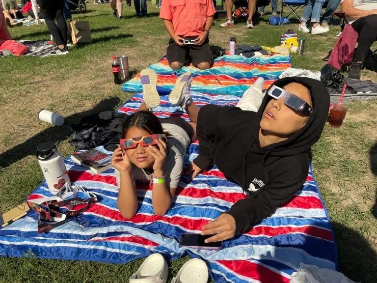 Two children sit on a blanket wearing eclipse glasses while waiting for the event to unfold.