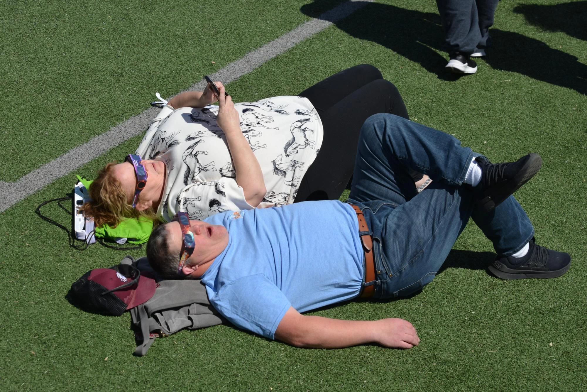 couple lying on field watching solar eclipse with glasses outdoor event — people viewing the eclipse with protective glasses