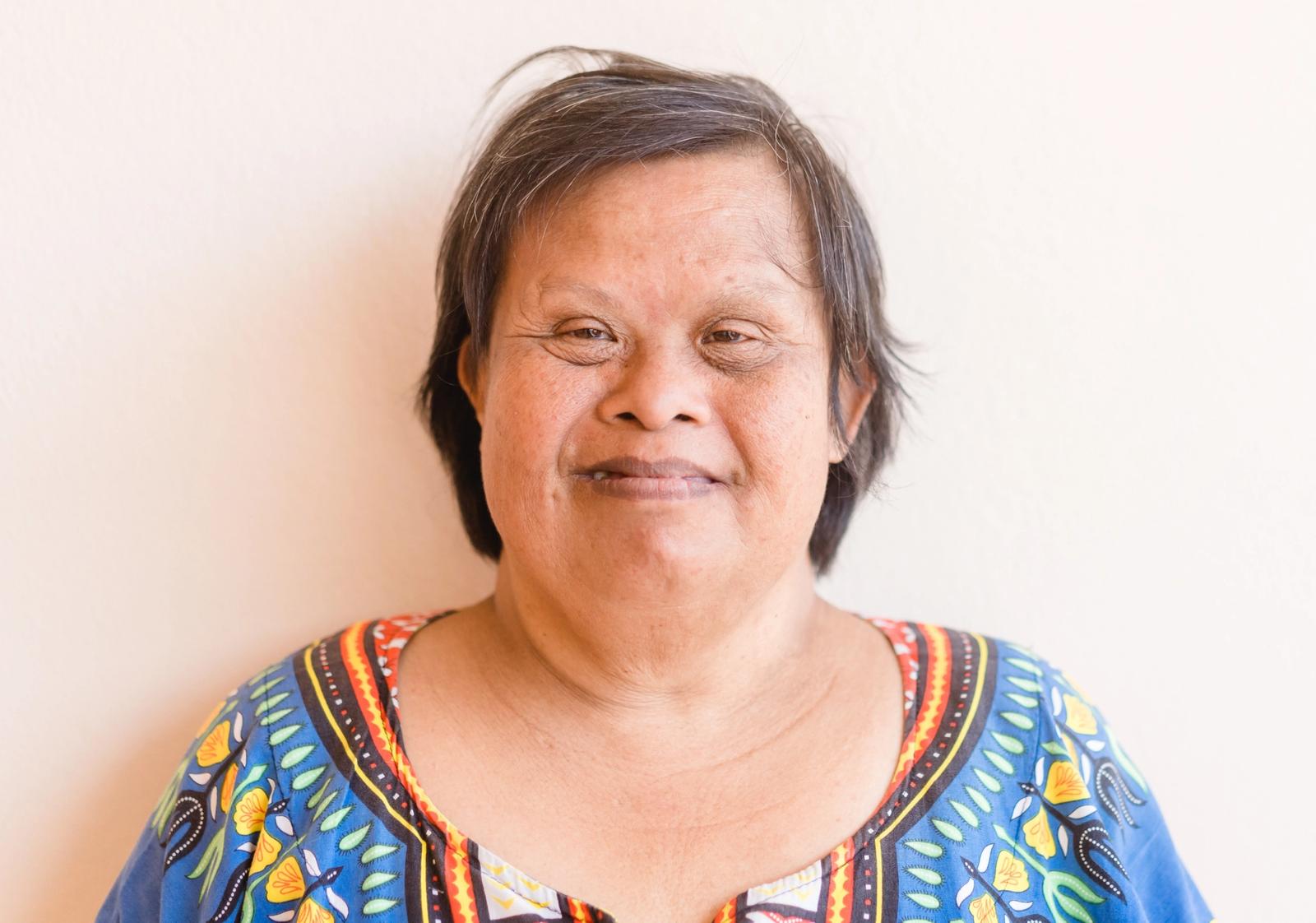 A mature woman with Down syndrome standing against a wall and smiling