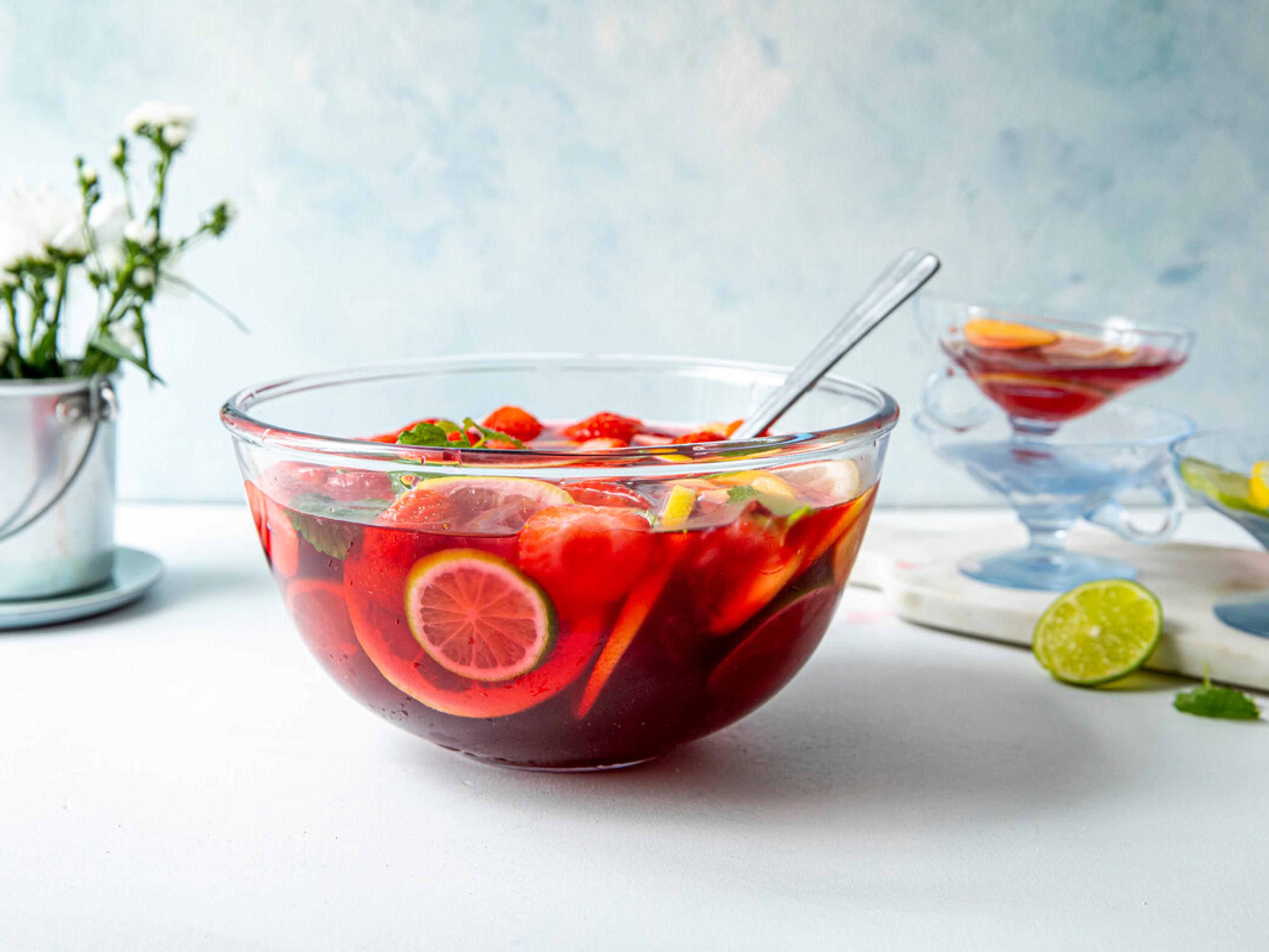 Stor bolle med rød sommerdrikk fylt med jordbær, sitrusfrukter og mynte. Drikken serveres med øse, og et glass med samme drikk står i bakgrunnen.