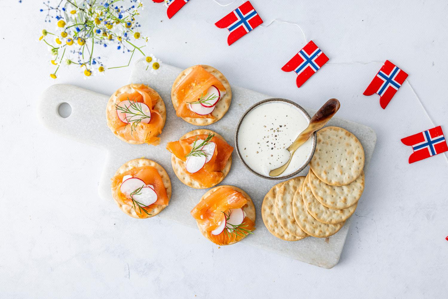 Kjeks toppet med røkelaks, dill og reddik servert på et fat sammen med rømme og norske flagg som dekorasjon.