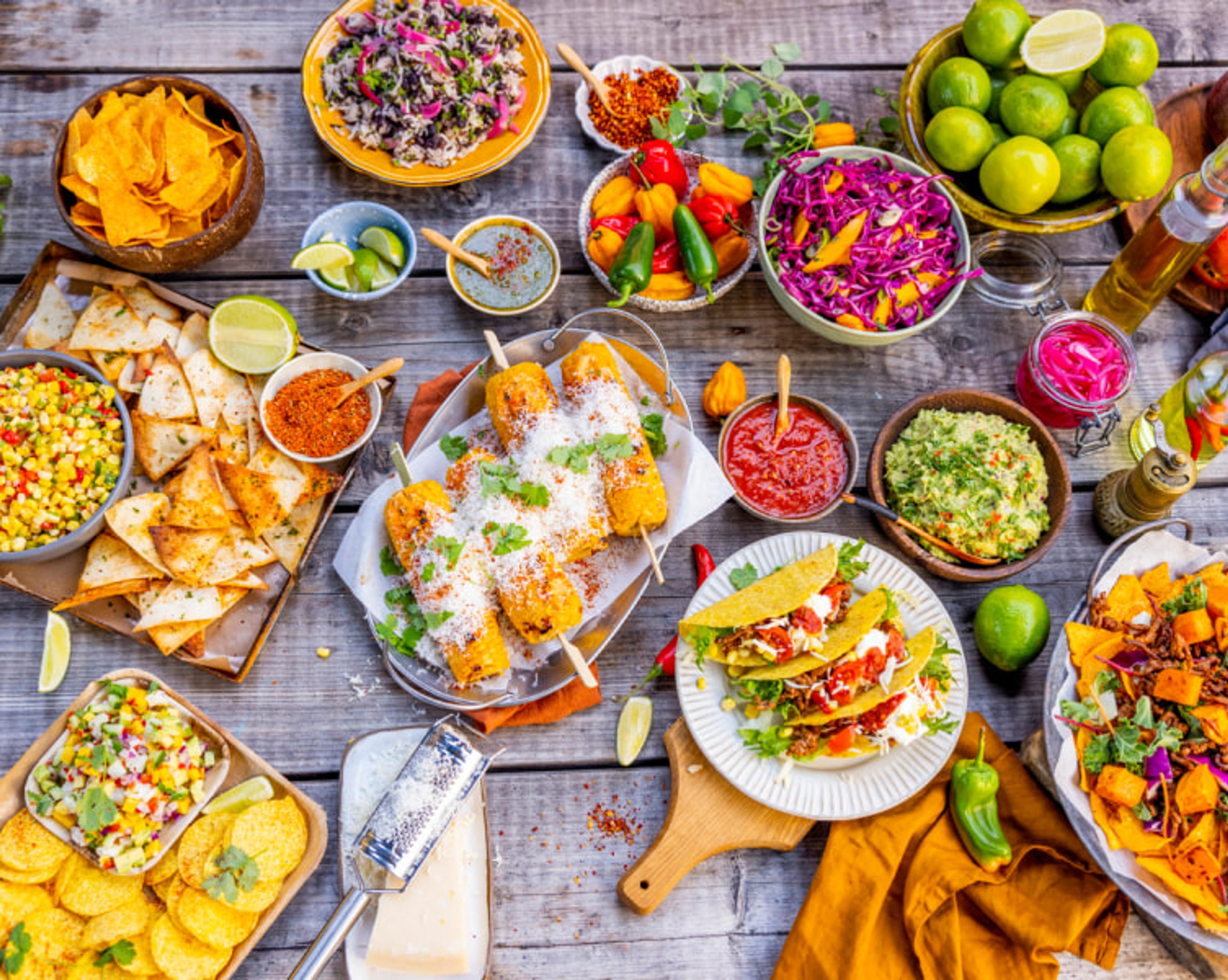 Fargerikt texmex-bord med tacos, grillede maiskolber, tortillachips, guacamole, salsa, salater, lime og ulike grønnsaker. Rettene er servert på et rustikt trebord med mange små tilbehørsskåler.