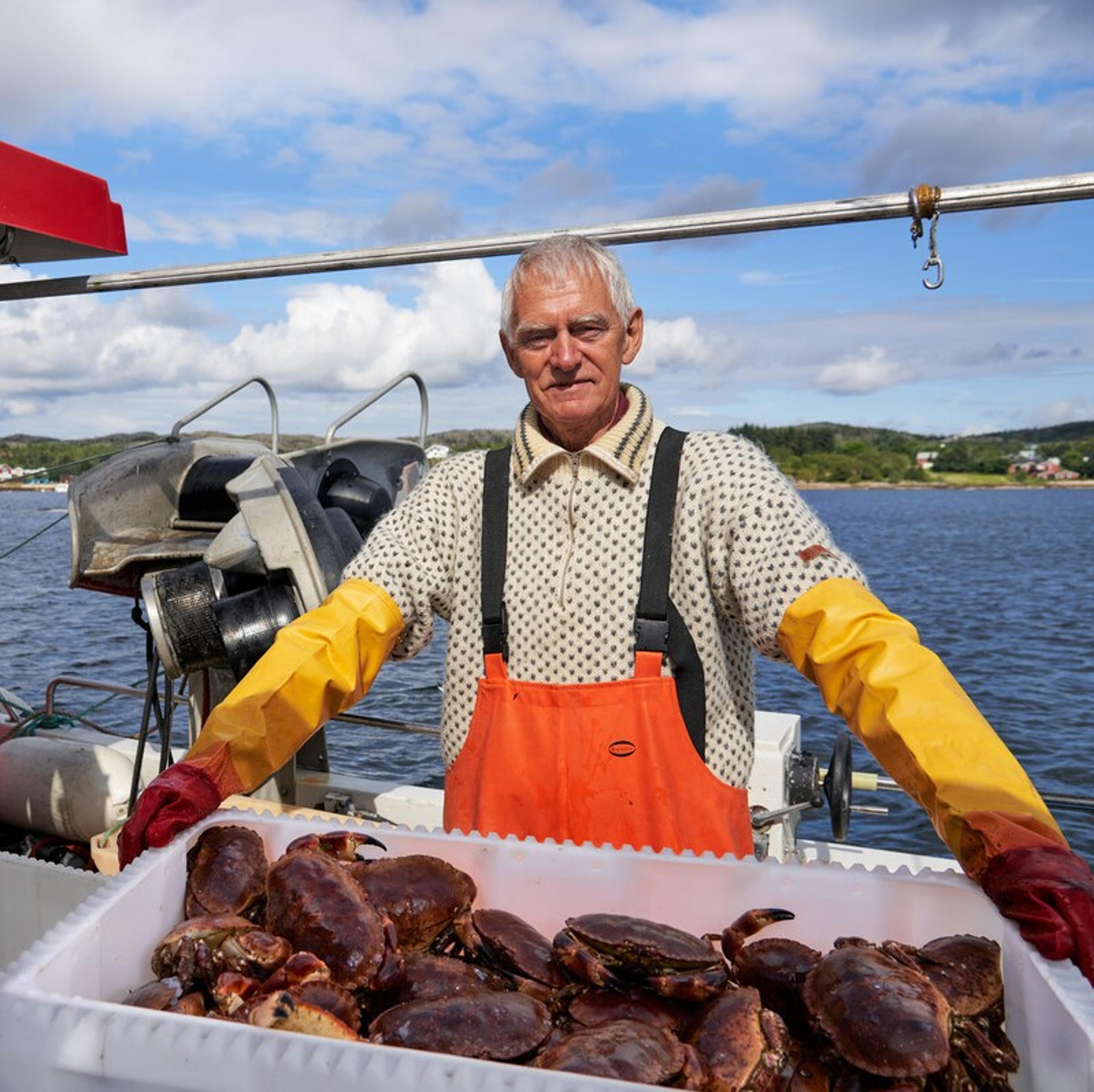 Person på fiskebåt står bak en kasse fylt med krabber. Personen har på seg oljehyre og gule hansker, og havet og kystlandskapet ses i bakgrunnen.