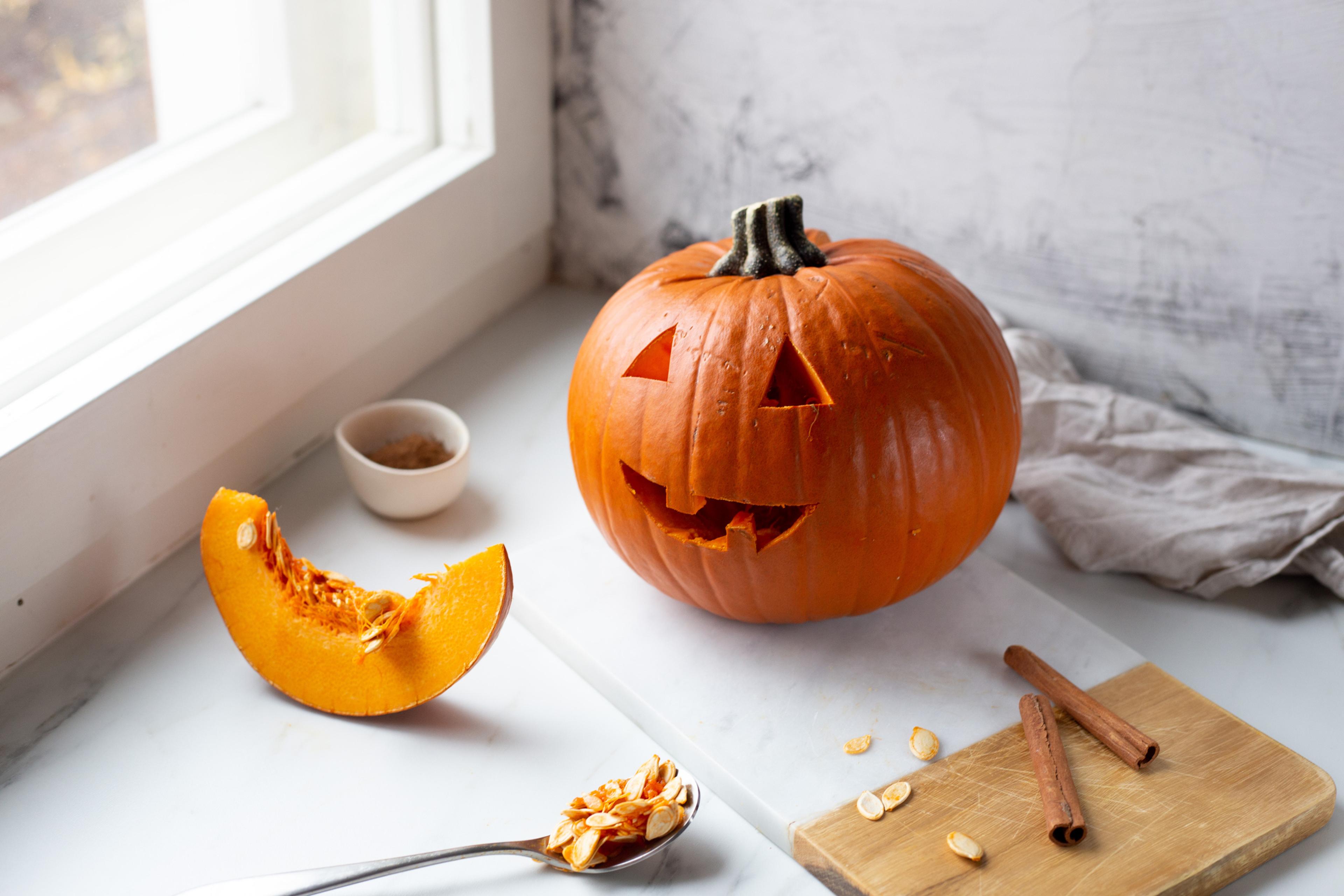 Utskåret Halloween-gresskar med trekantede øyne og smilende munn ved siden av et stykke gresskar og gresskarfrø.