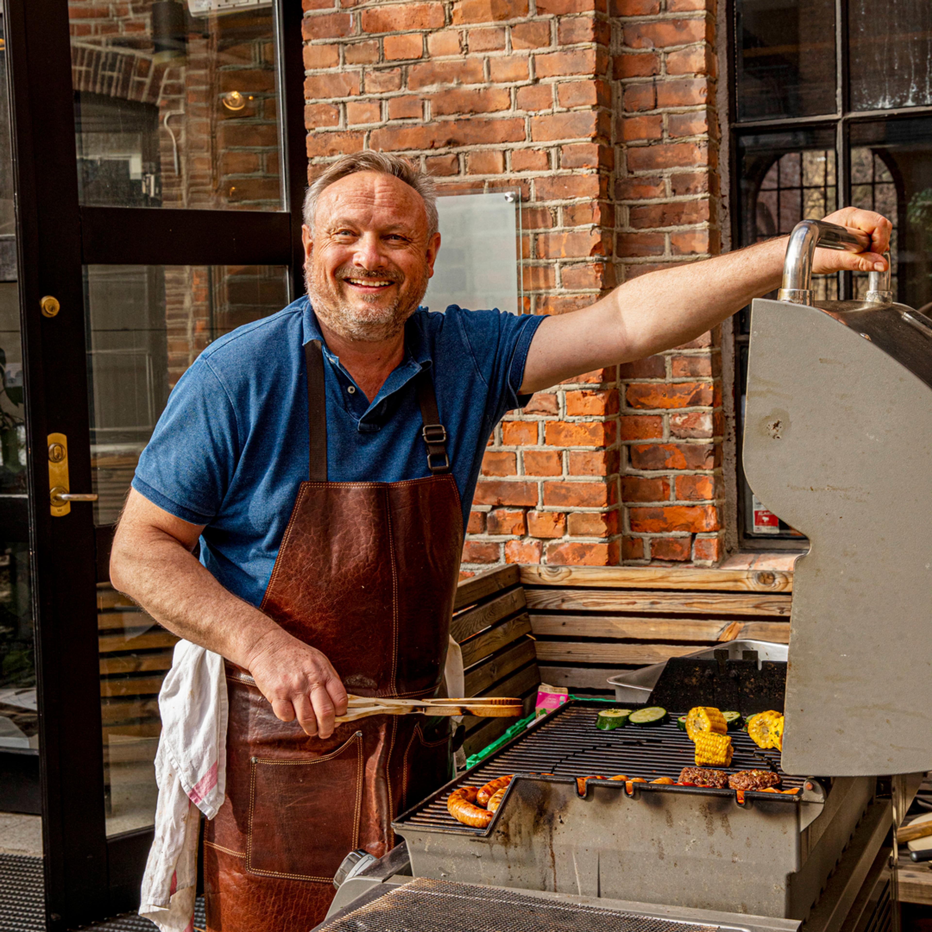 Bent Stiansen i skinnforkle smiler mens han snur maiskolber, pølser og grillburgere på en gassgrill foran en murvegg