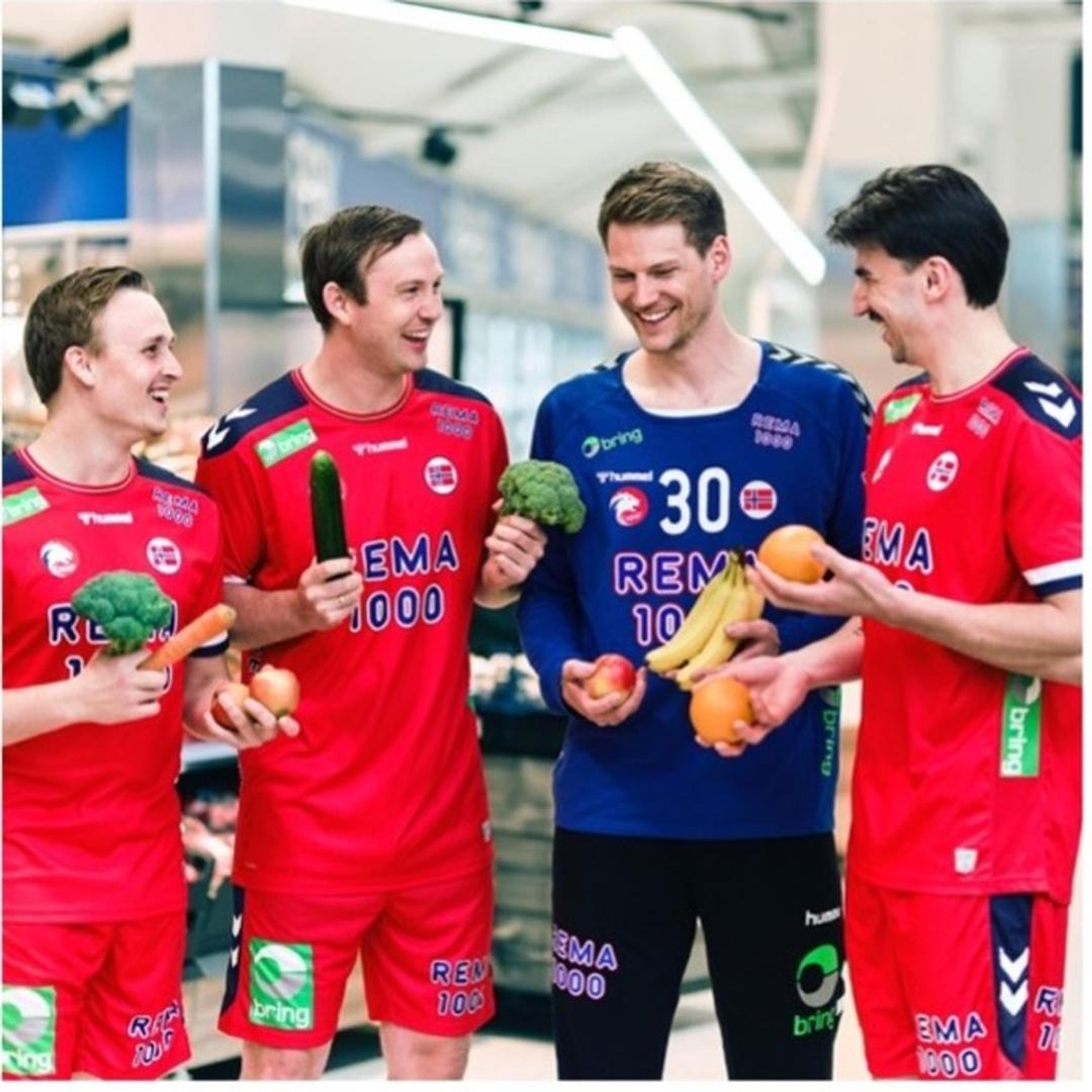 Fire håndballspillere i REMA 1000-drakter står i en dagligvarebutikk og holder ulike frukter og grønnsaker, blant annet brokkoli, gulrot, agurk, bananer og appelsiner. Frukt- og grøntavdelingen vises i bakgrunnen.