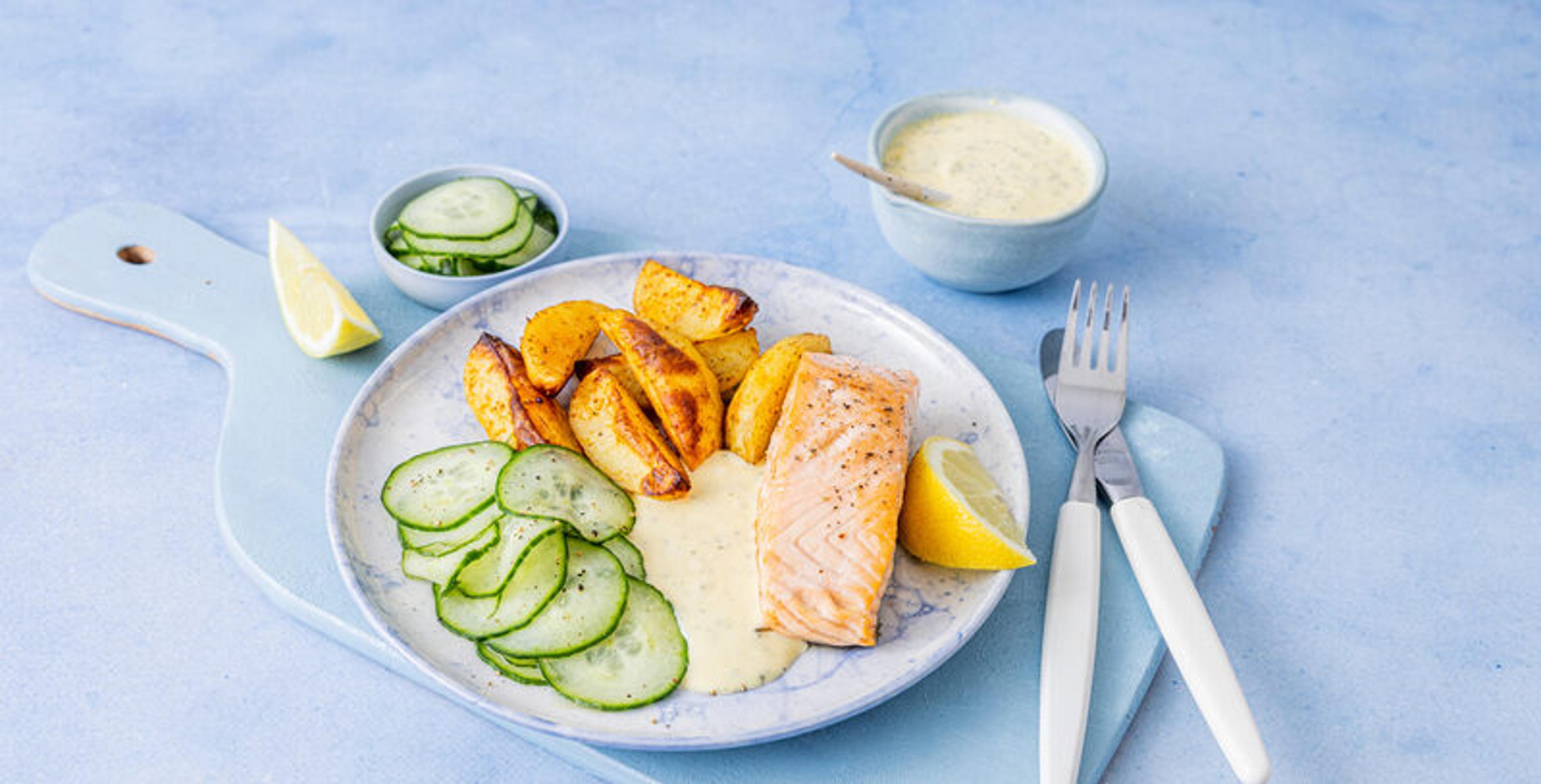 Stekt laksefilet servert med ovnsbakte potetbåter, agurksalat, kremet saus og sitronbåter på en lys tallerken.