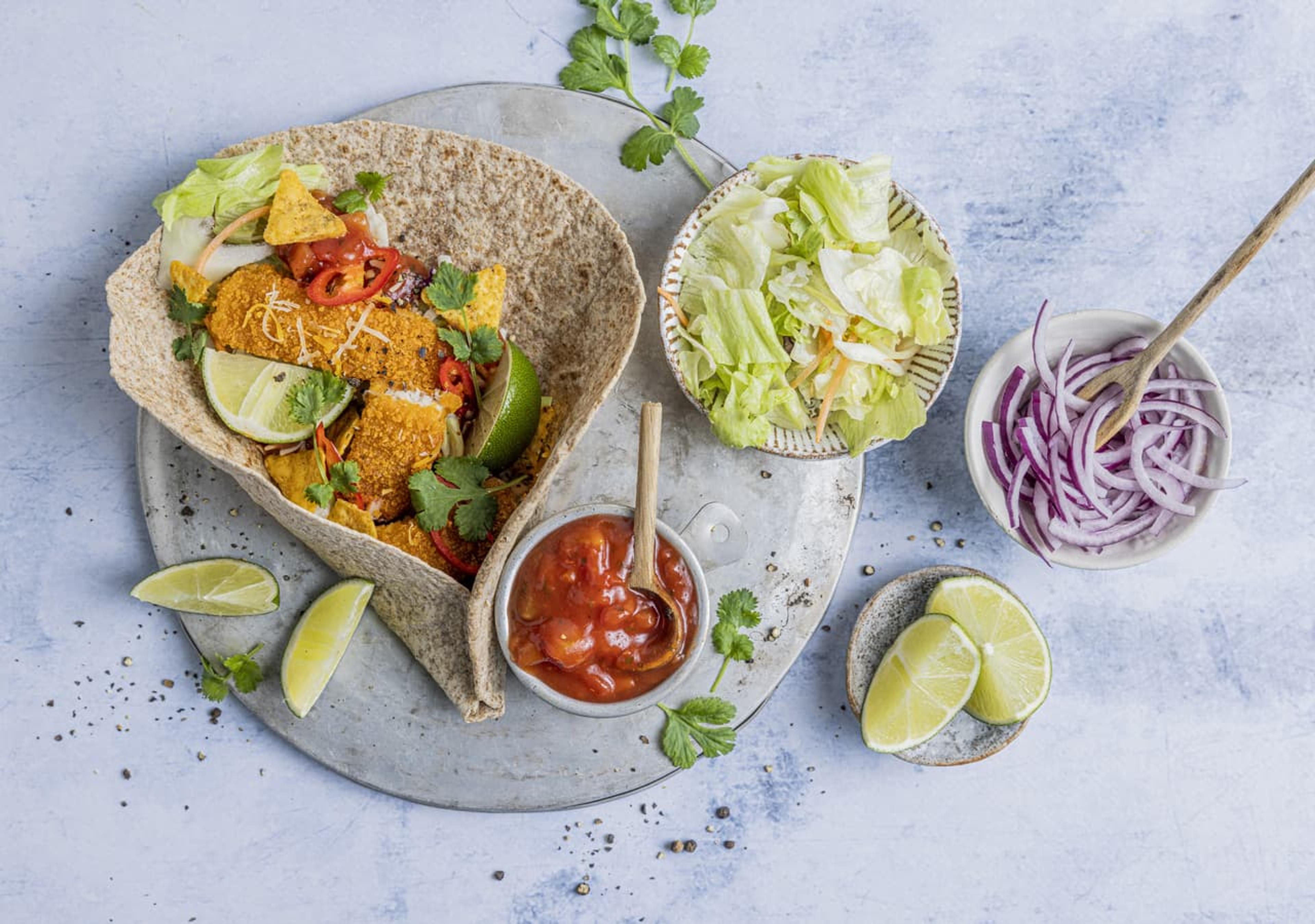fisketaco som består av lefse, fiskepinner i biter, lime, chili, rødløk og salsa