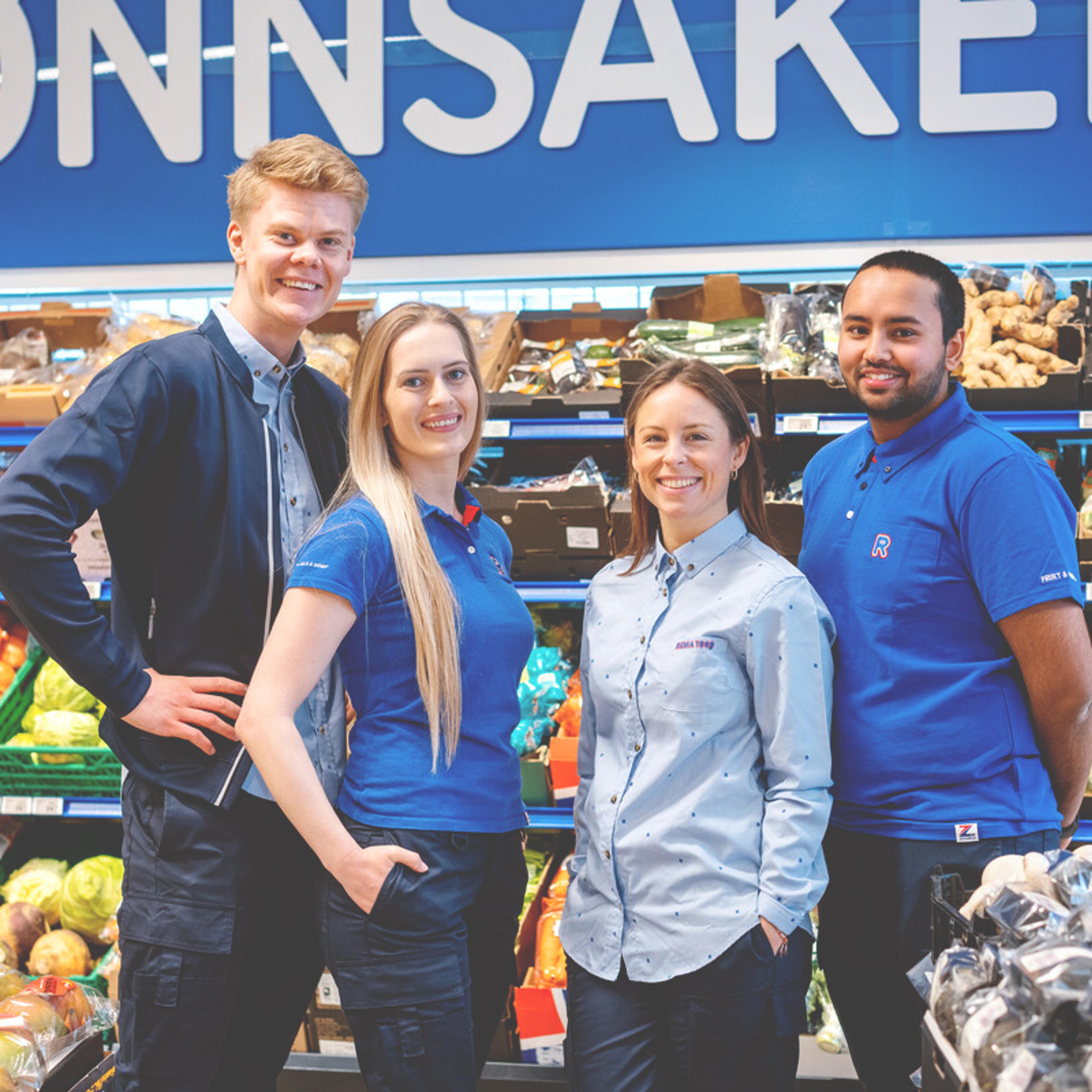Fire butikkmedarbeidere står foran frukt- og grøntavdelingen i en dagligvarebutikk. Hyller med ferske grønnsaker vises i bakgrunnen, og et blått skilt med teksten «GRØNNSAKER» henger over avdelingen.