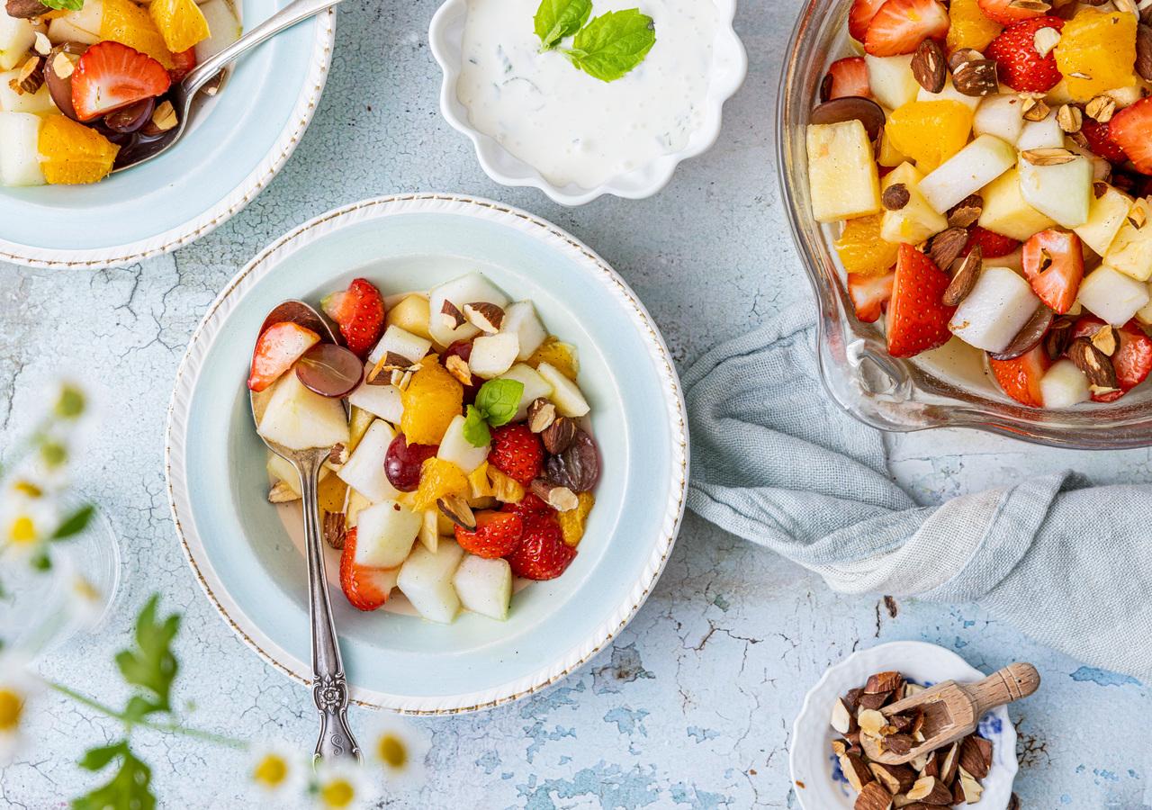 Fruktsalat med jordbær, melon, appelsin, druer og hakkede mandler servert i skål.