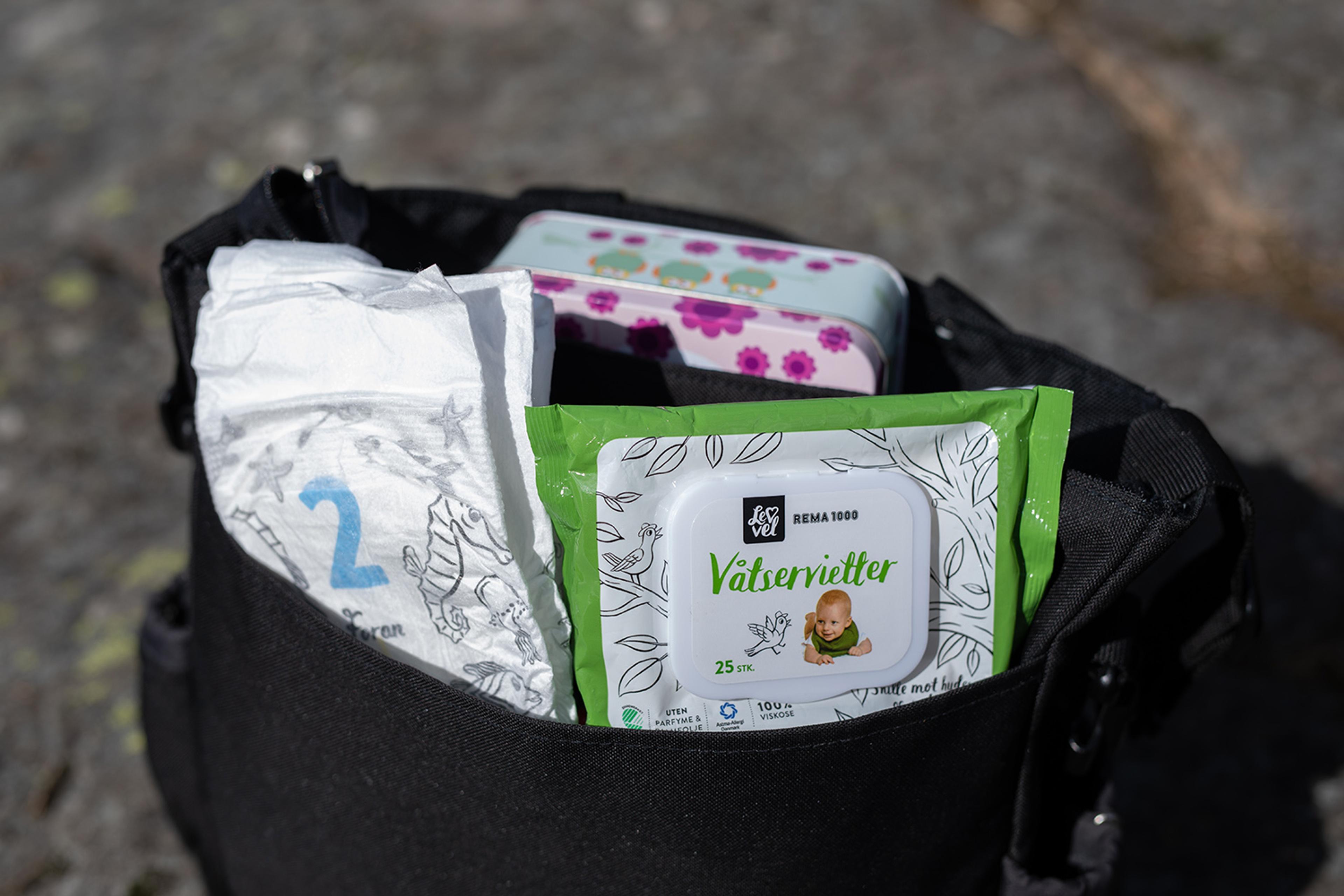 Åpen stelleveske med bleie, våtservietter og matboks. Produktene ligger lett tilgjengelig i vesken og er fotografert utendørs på steingrunn.