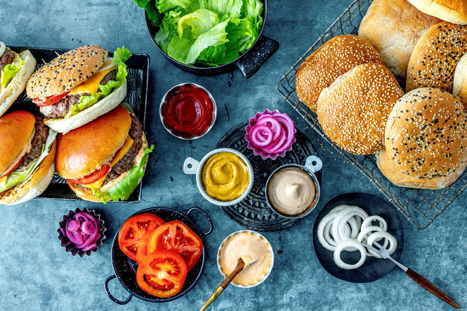 En blå bordplate dekket med deilige burgere, burgerbrød og årets tilbehør som dipper og dressinger.