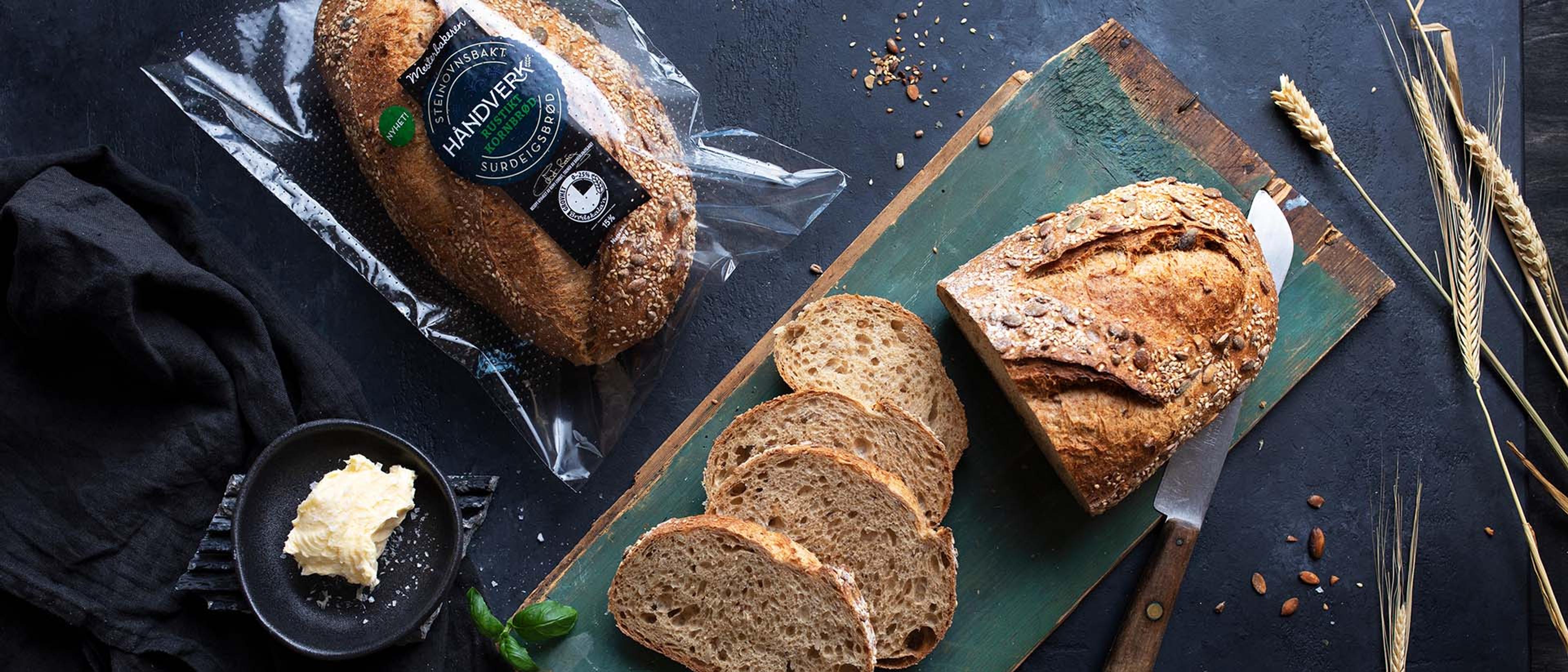 Oppskåret håndverksbrød med frø og korn på en skjærefjøl, servert med smør. Et helt brød i emballasje ligger ved siden av, omgitt av kornaks og frø på mørk bakgrunn.