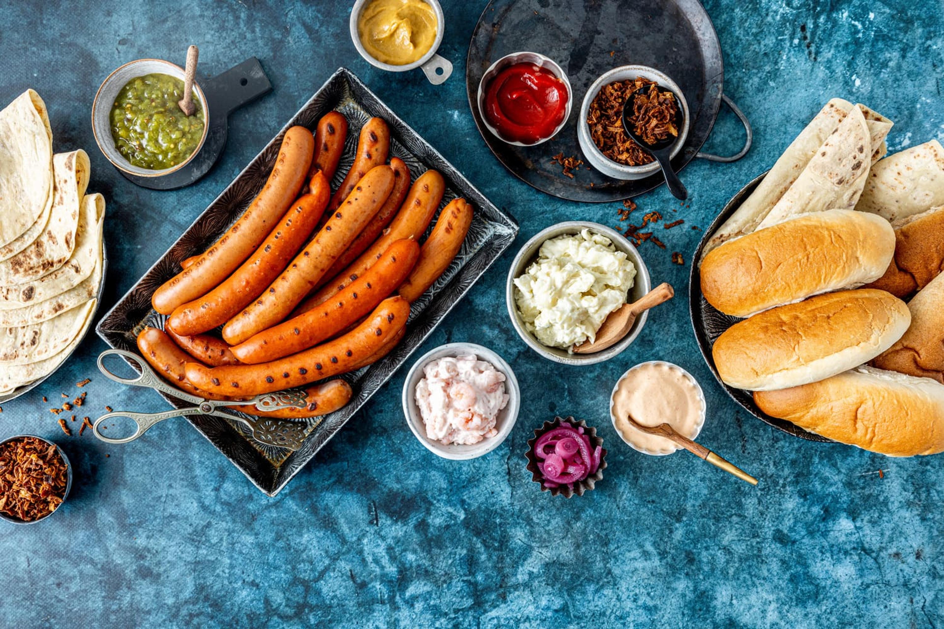 En blå bordplate dekket med smakfulle grilla pølser med tilbehør som pølsebrød, lomper og diverse av årets nye dipper.
