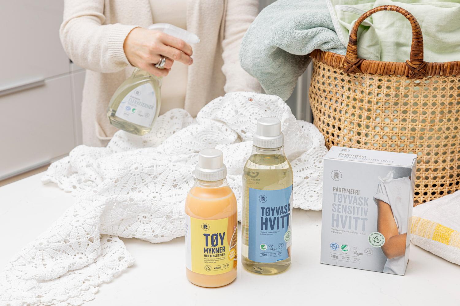 Vaskeromsmiljø med hvite tekstiler, vaskekurv og vaskemidler fra REMA 1000. Produktene inkluderer tøymykner, flytende vaskemiddel for hvitt tøy og parfymefri vaskepulver, mens en person holder en flekkfjerner over tekstilene.