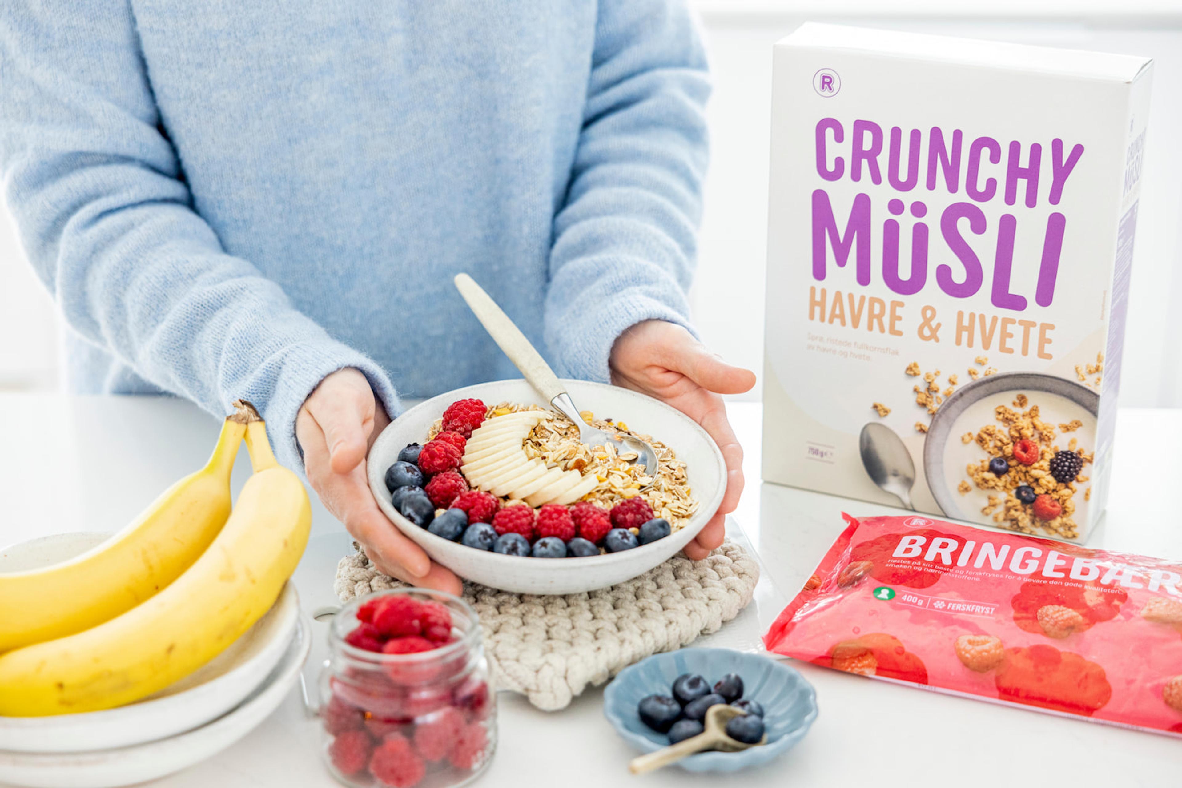 Skål med crunchy müsli toppet med banan, bringebær og blåbær, ved siden av müsli, bringebær og bananer.