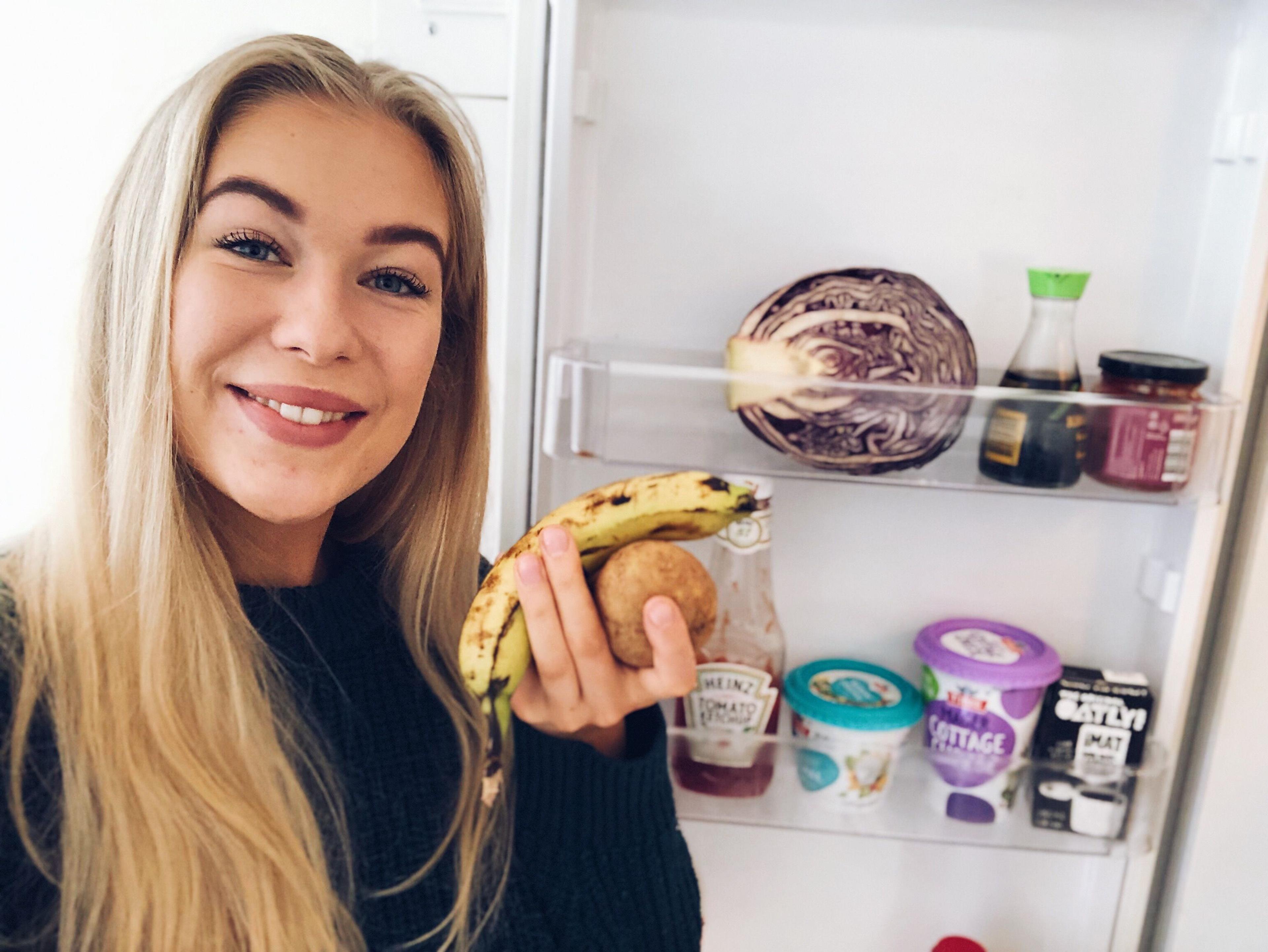 Person holder en banan foran et åpent kjøleskap med ulike matvarer.