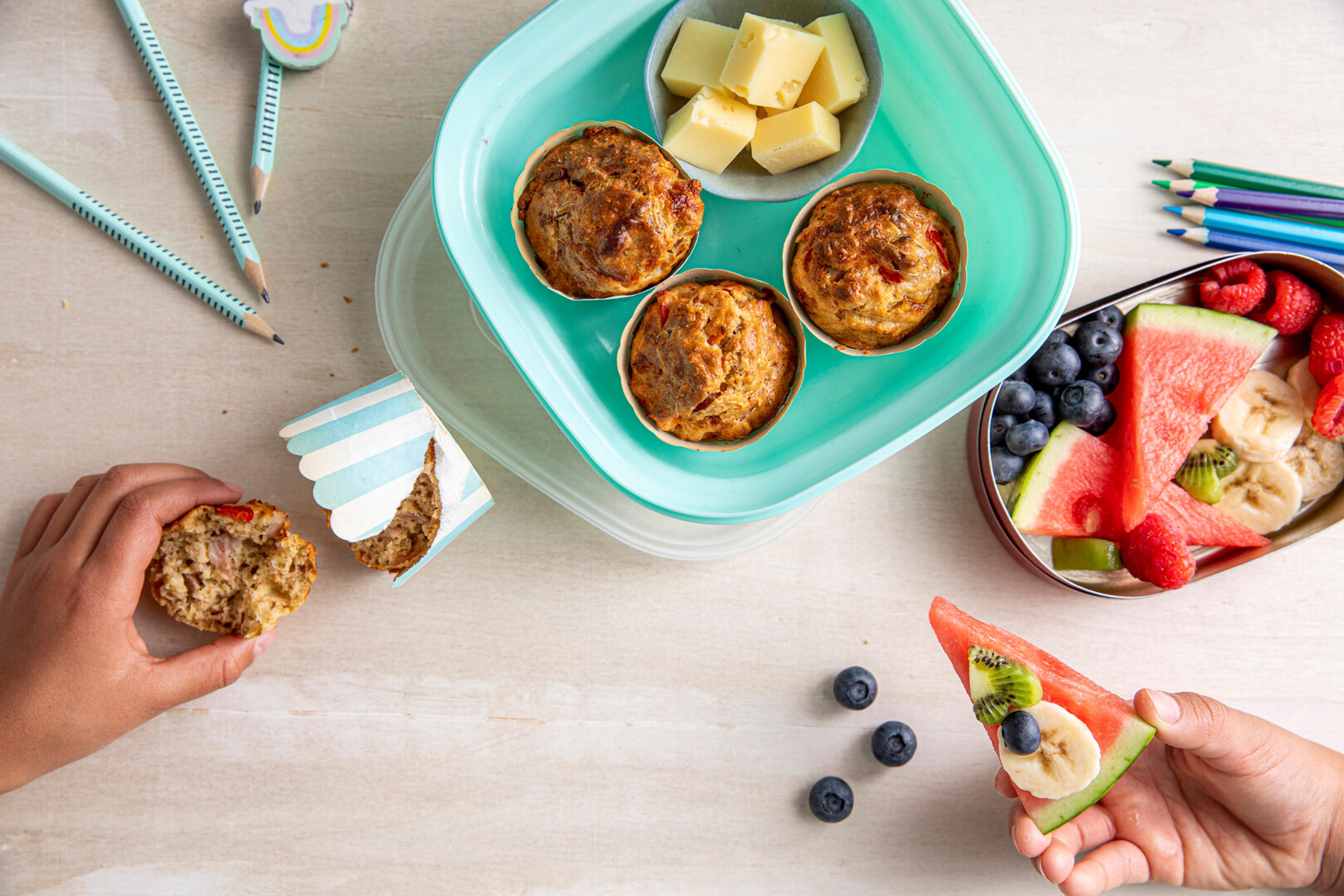 Matpakke med grove muffins, ostebiter og oppskåret frukt og bær. To hender holder henholdsvis en muffins og en vannmelonskive toppet med banan, kiwi og blåbær.