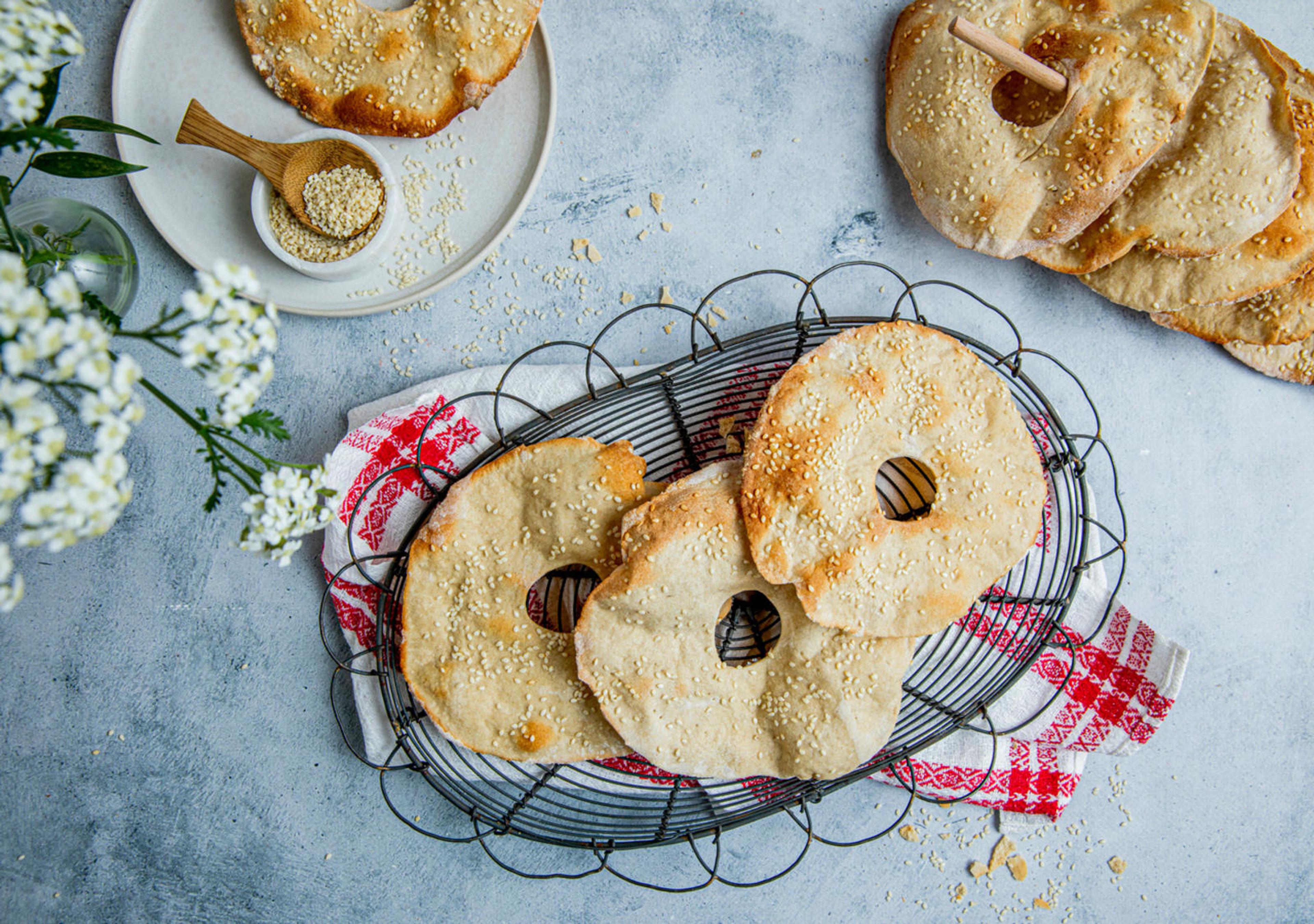 Hjemmebakte sesambrød med hull i midten ligger på rist og tallerkener. Brødene er toppet med sesamfrø og servert på et lyst underlag.