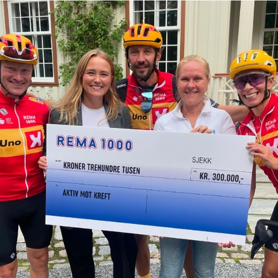Fem personer står samlet utendørs og holder en stor gave-/sjekkplakat fra REMA 1000 på 300 000 kroner til Aktiv mot kreft. To personer i sykkelbekledning og hjelm står på hver side av gruppen, mens et hus og grønn vegetasjon ses i bakgrunnen.
