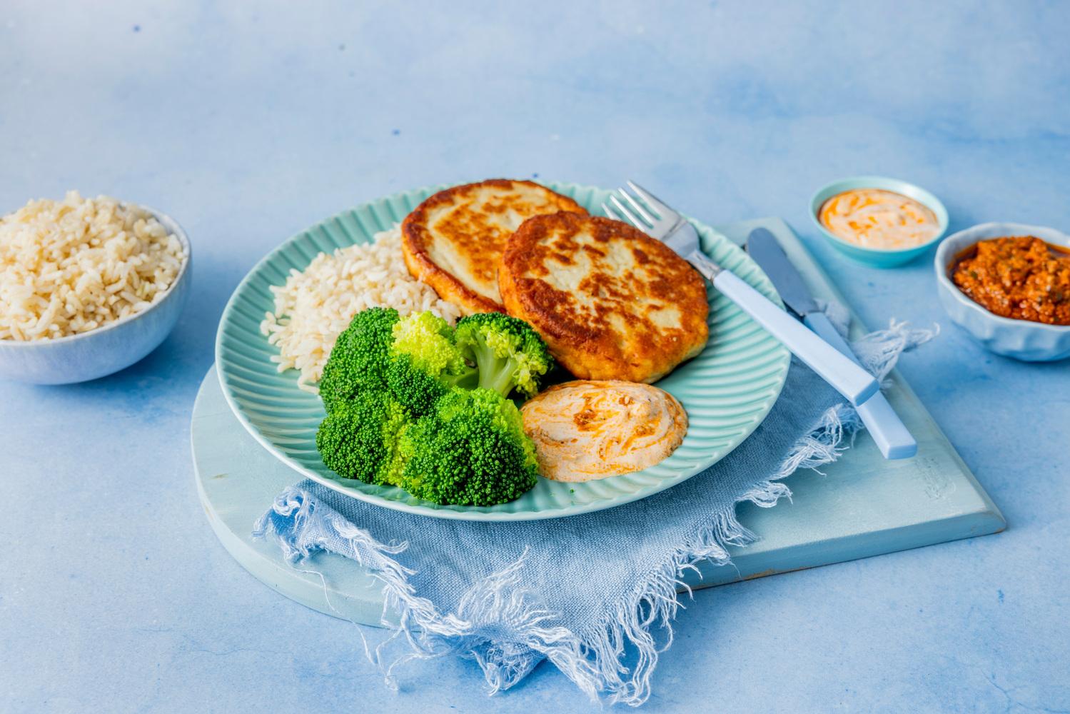 Panerte fiskekaker servert med ris, brokkoli og saus på en tallerken. Ved siden av står små skåler med ris og tilbehør.