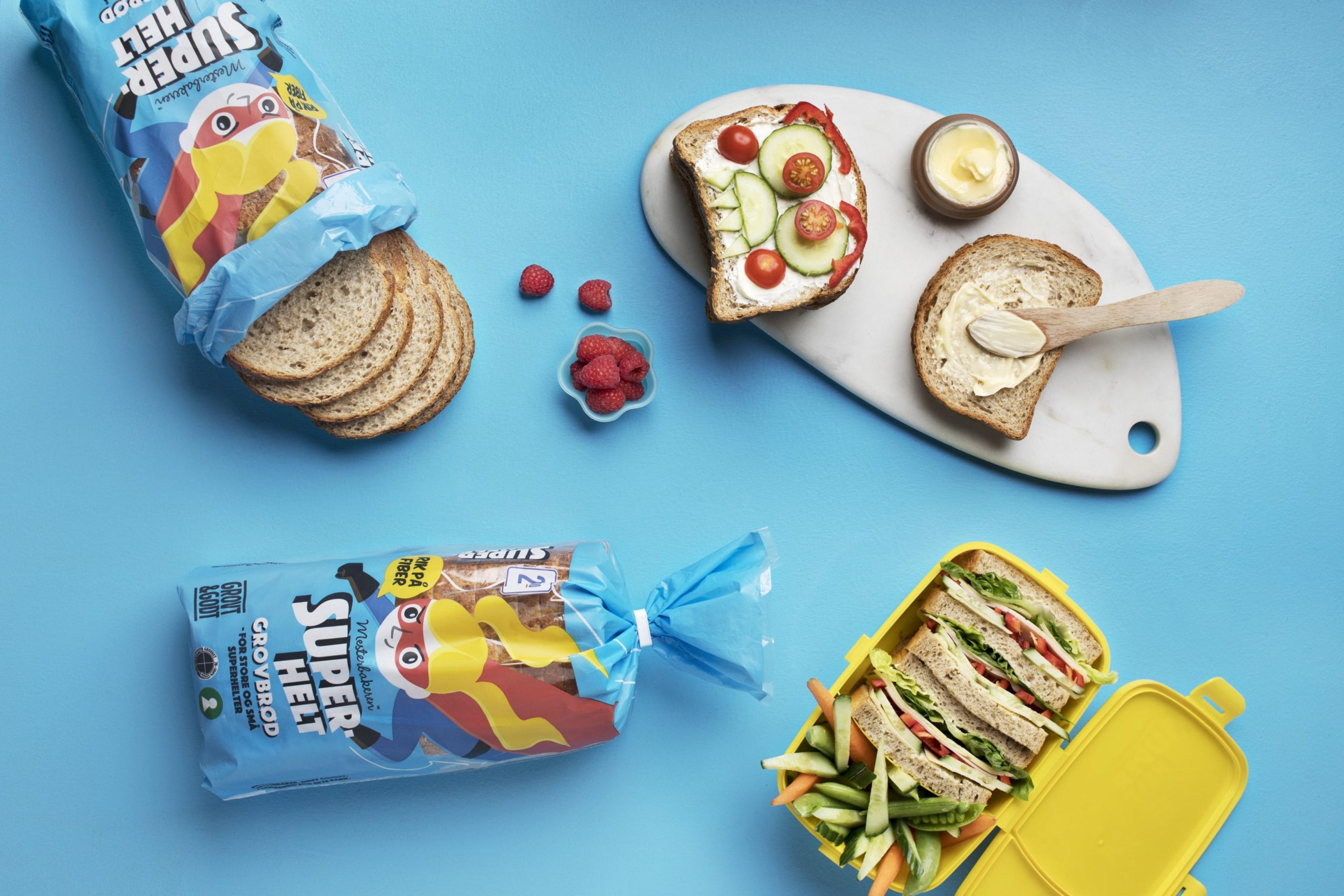 Skiver av superheltbrød med pålegg og grønnsaker på blå bakgrunn. To brødposer, åpne smørbrød med agurk og tomat, bringebær og matboks med sandwich og oppskårne grønnsaker.