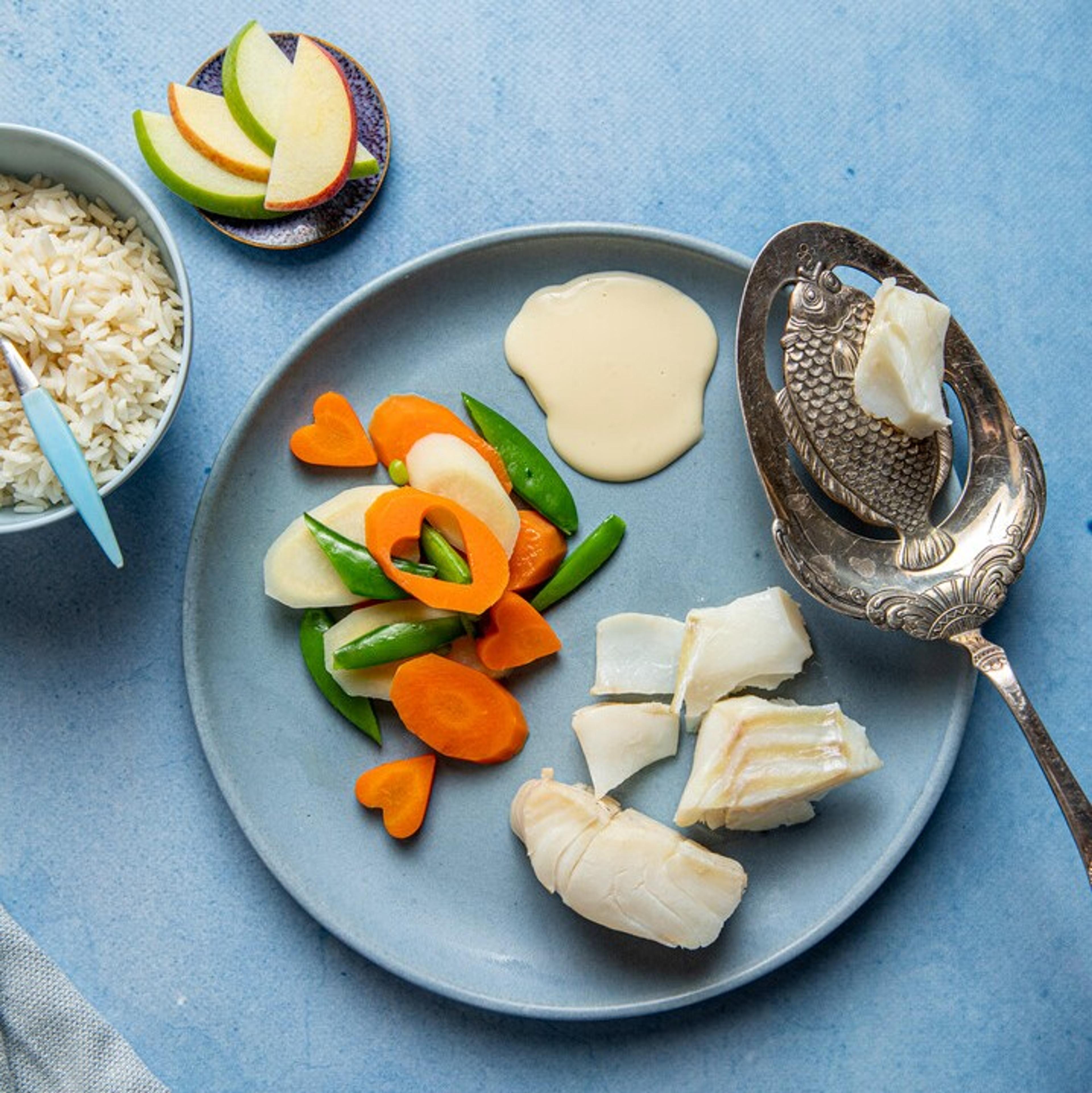Tallerken med kokt hvit fisk, grønnsaker formet som hjerter, hvit saus og epleskiver. Ved siden av står en skål med ris og et serveringsfat med smør.