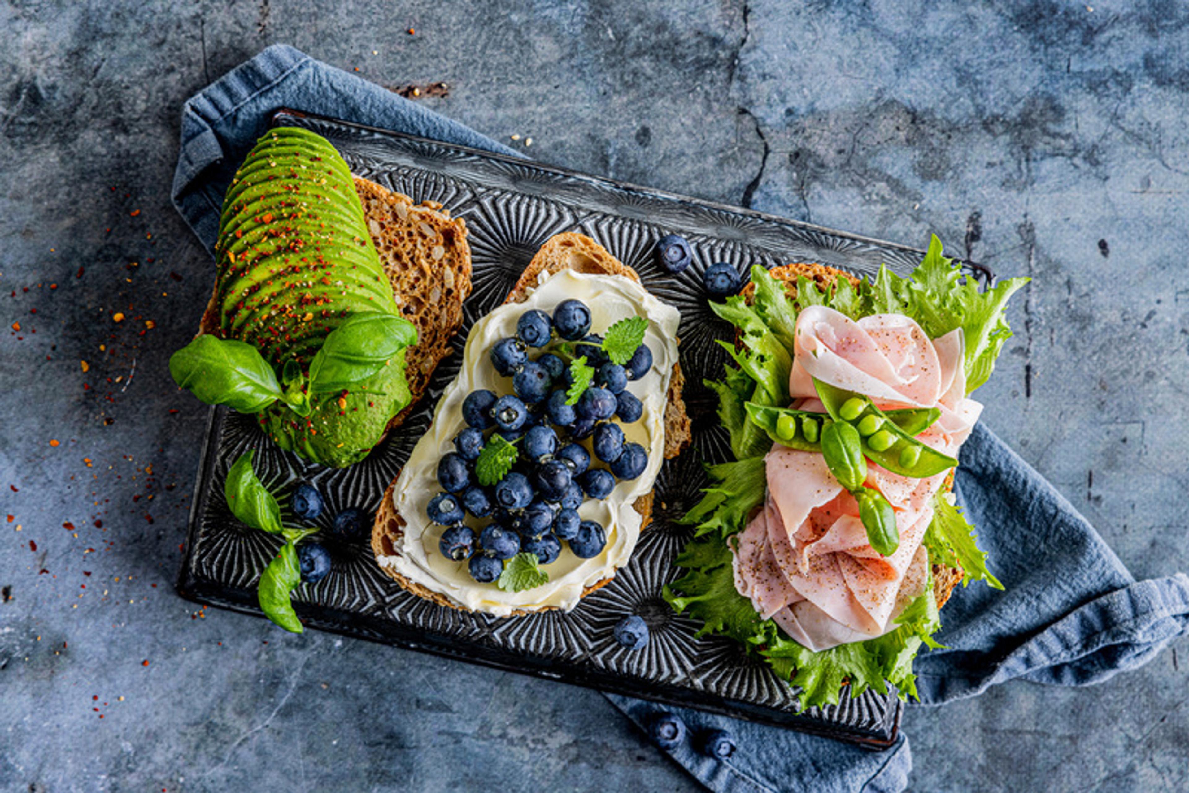 Tre åpne brødskiver med ulikt pålegg på et fat. Én med skivet avokado, én med kremost og blåbær, og én med salat, skinke og sukkererter. Servert på mørk steinbakgrunn.