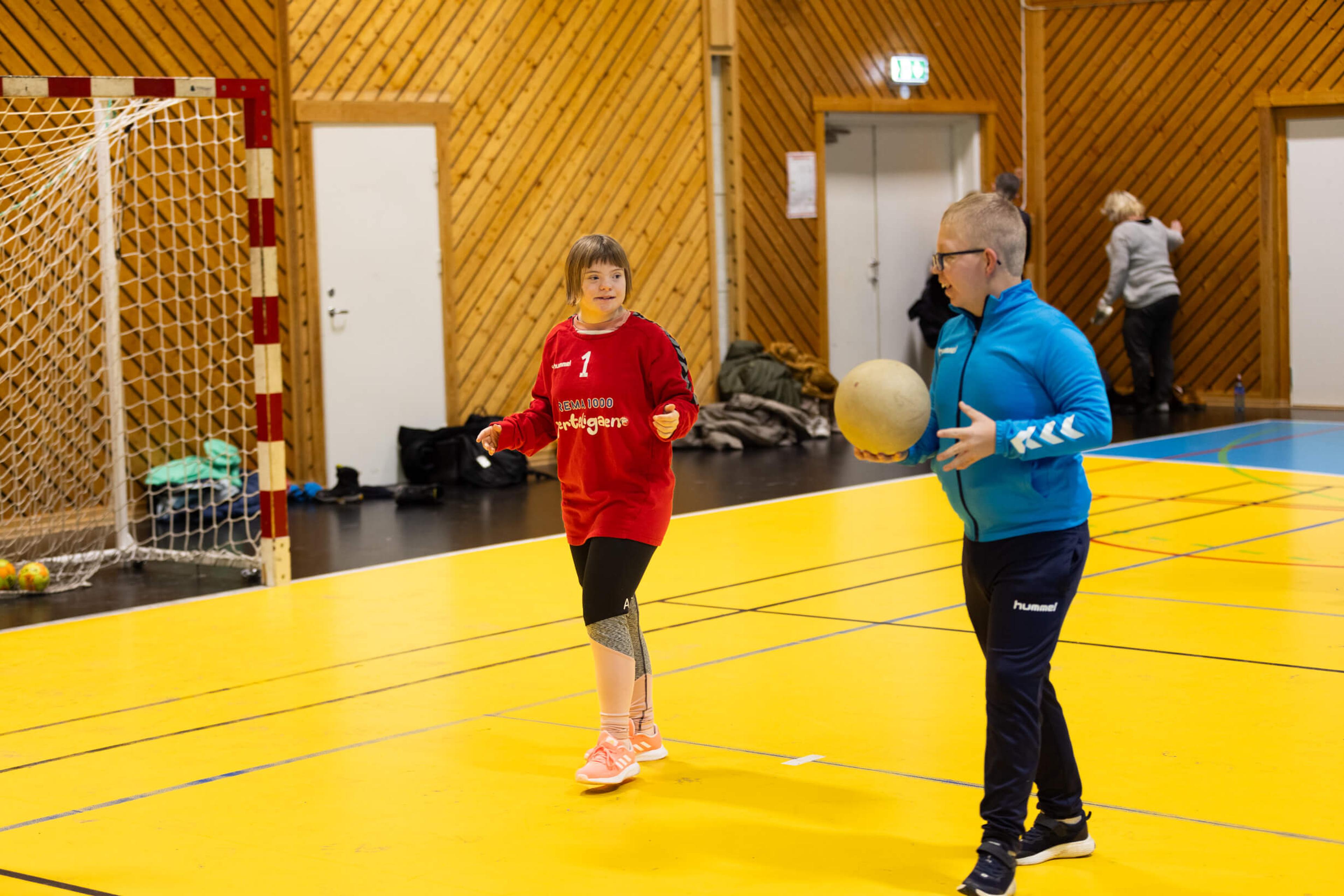 To deltakere i REMA 1000 Hjerteligaen spiller med ball i en idrettshall ved siden av et håndballmål.